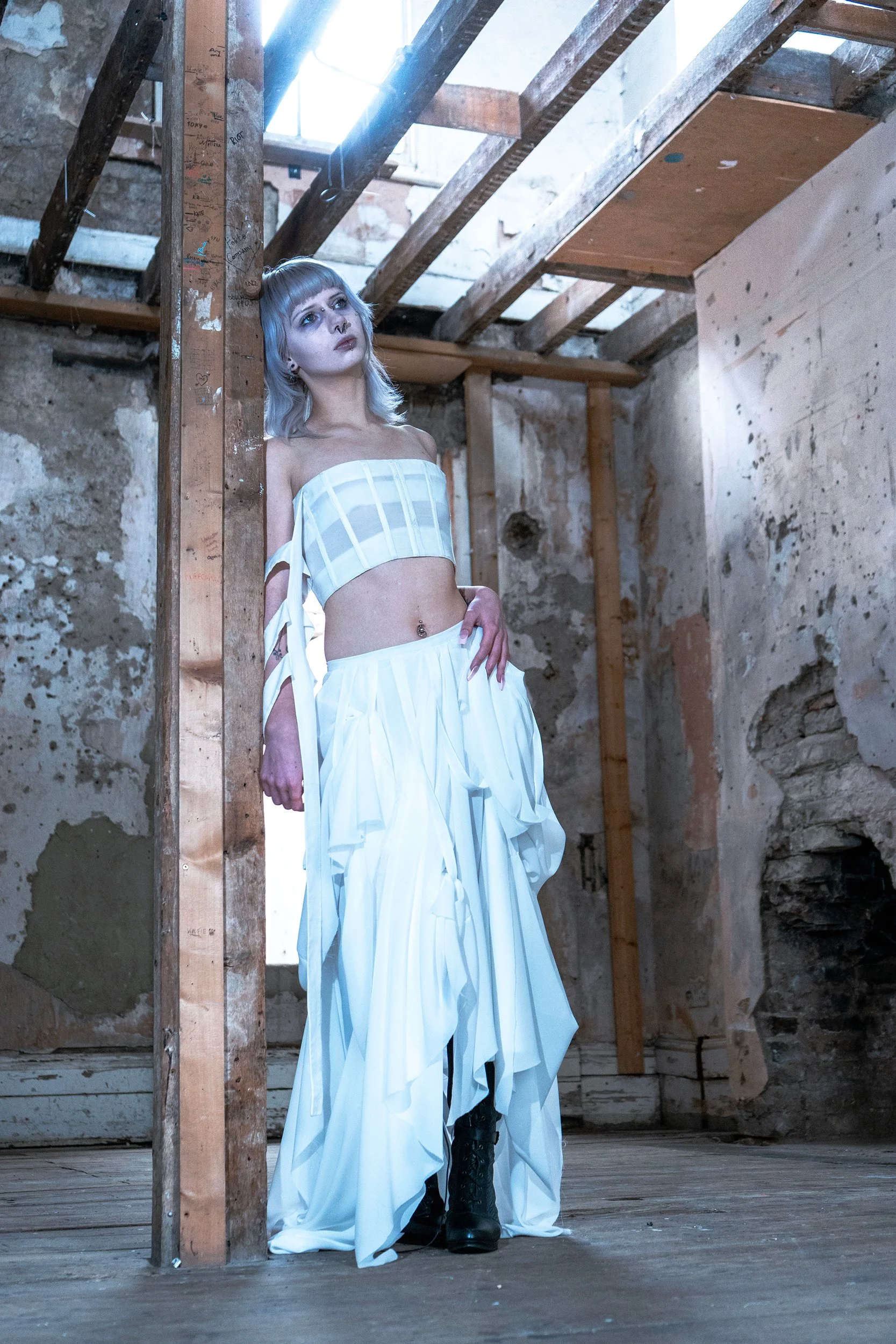 A woman with gray hair and makeup, wearing a strapless white top and flowing white skirt, standing in a rustic, unfinished room with exposed wooden beams and brick walls, with sunlight coming through a window.