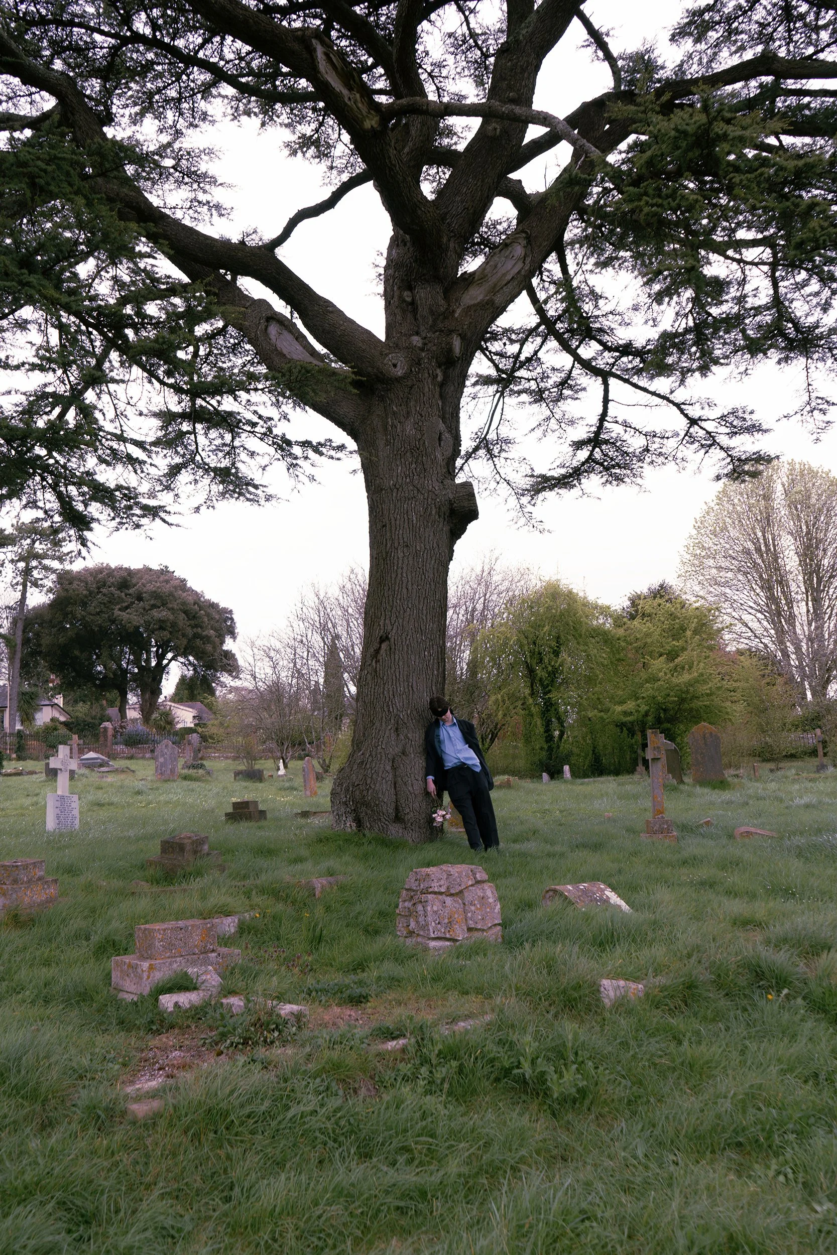 A person dressed in dark clothing leaning against a large tree in a graveyard.