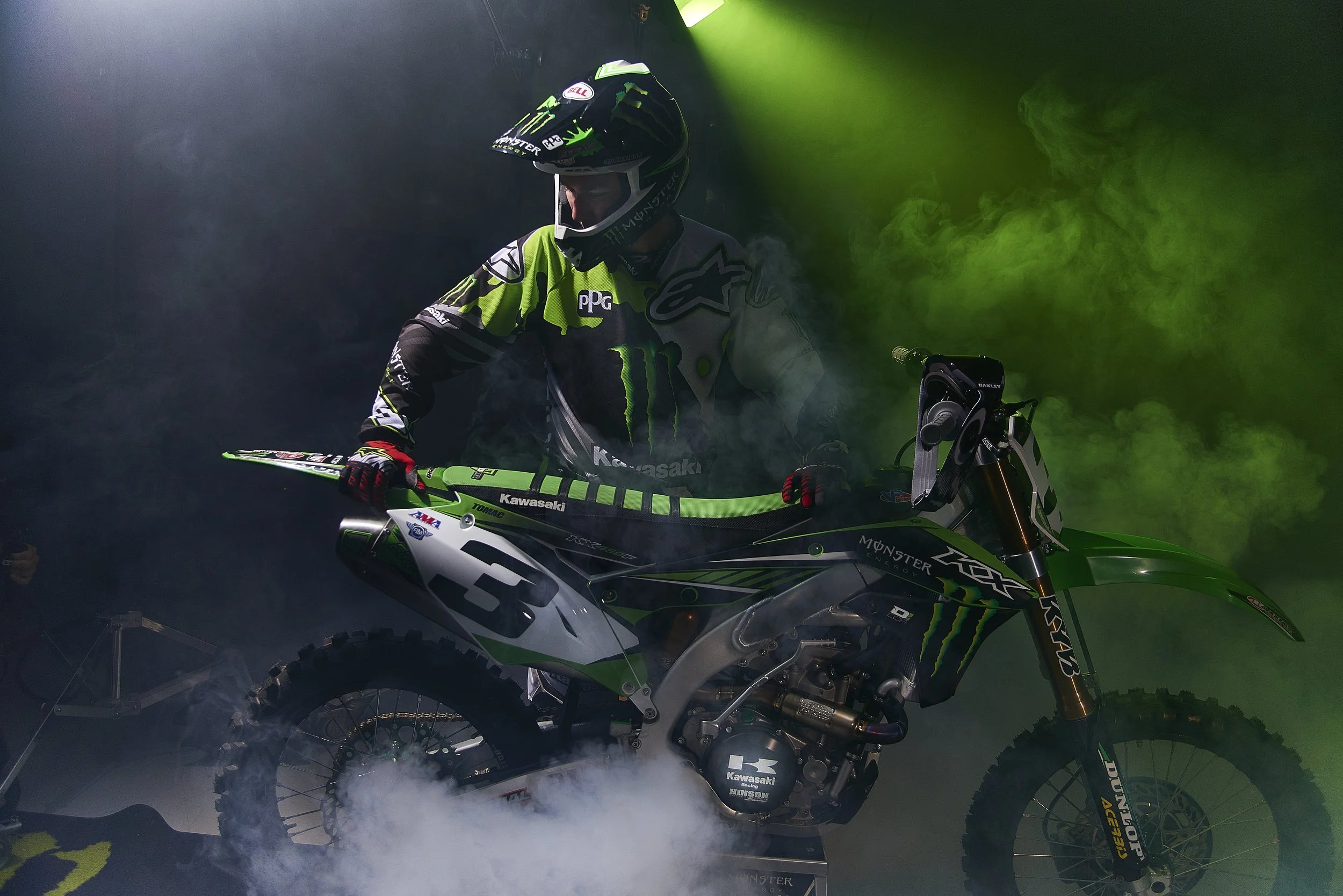 A professional motocross rider in racing gear, including a helmet and a green and black Monster Energy Kawasaki motorcycle, is surrounded by green smoke and fog.