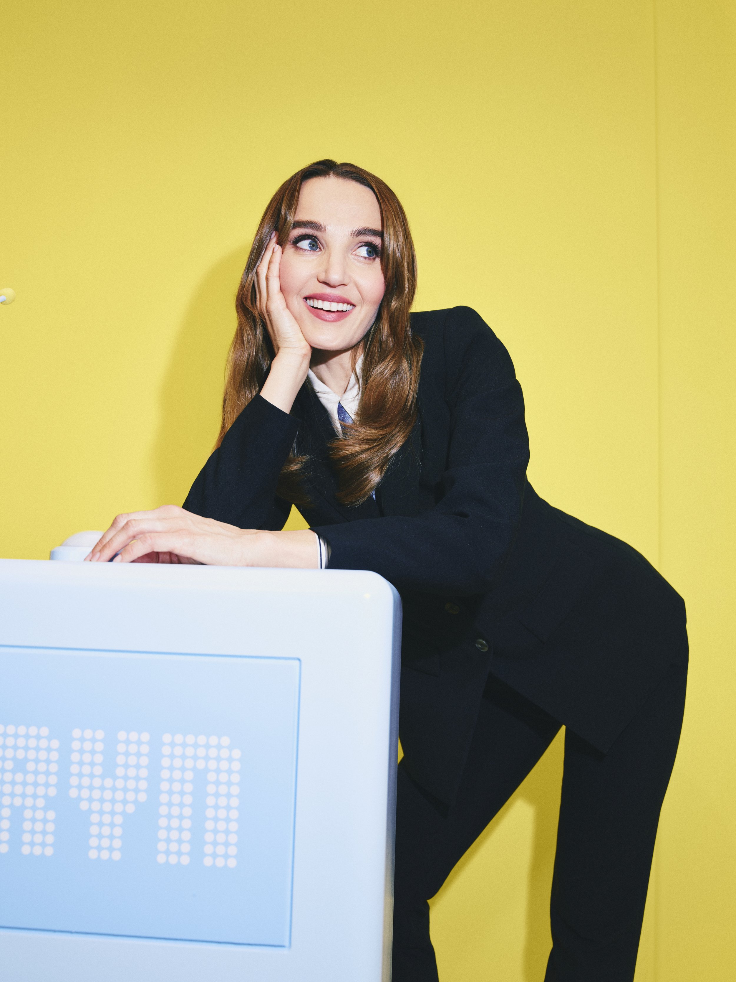 A woman with long brown hair and blue eyes, wearing a black blazer and white shirt, is smiling and resting her face on her hand indoors against a yellow wall, with a light blue computer monitor in front of her.