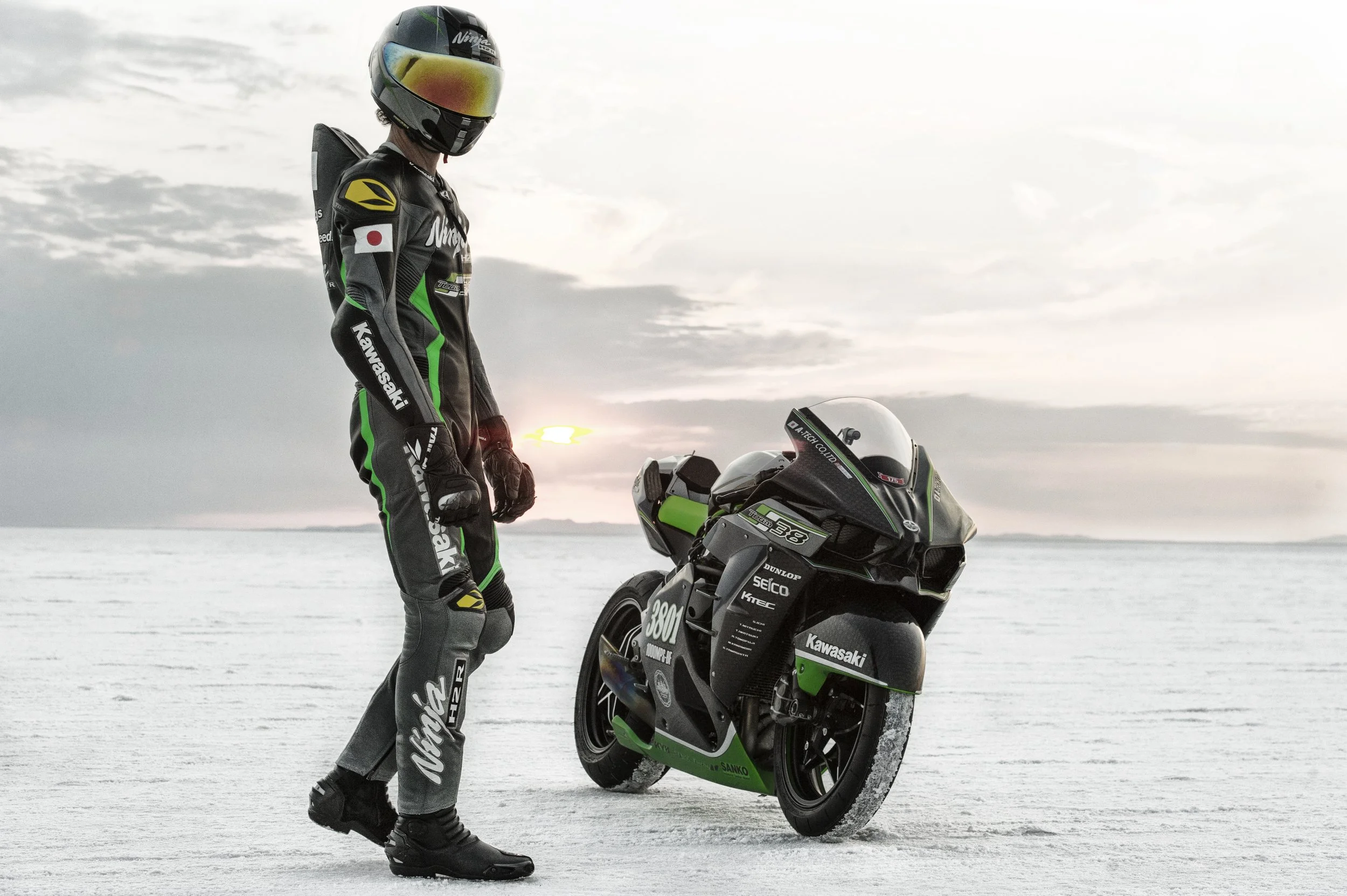 A person dressed in racing gear stands next to a green and black Kawasaki racing motorcycle on a salt flat or ice surface. The person wears a black helmet with a reflective visor, a black racing suit with green and yellow accents, and black boots. Th