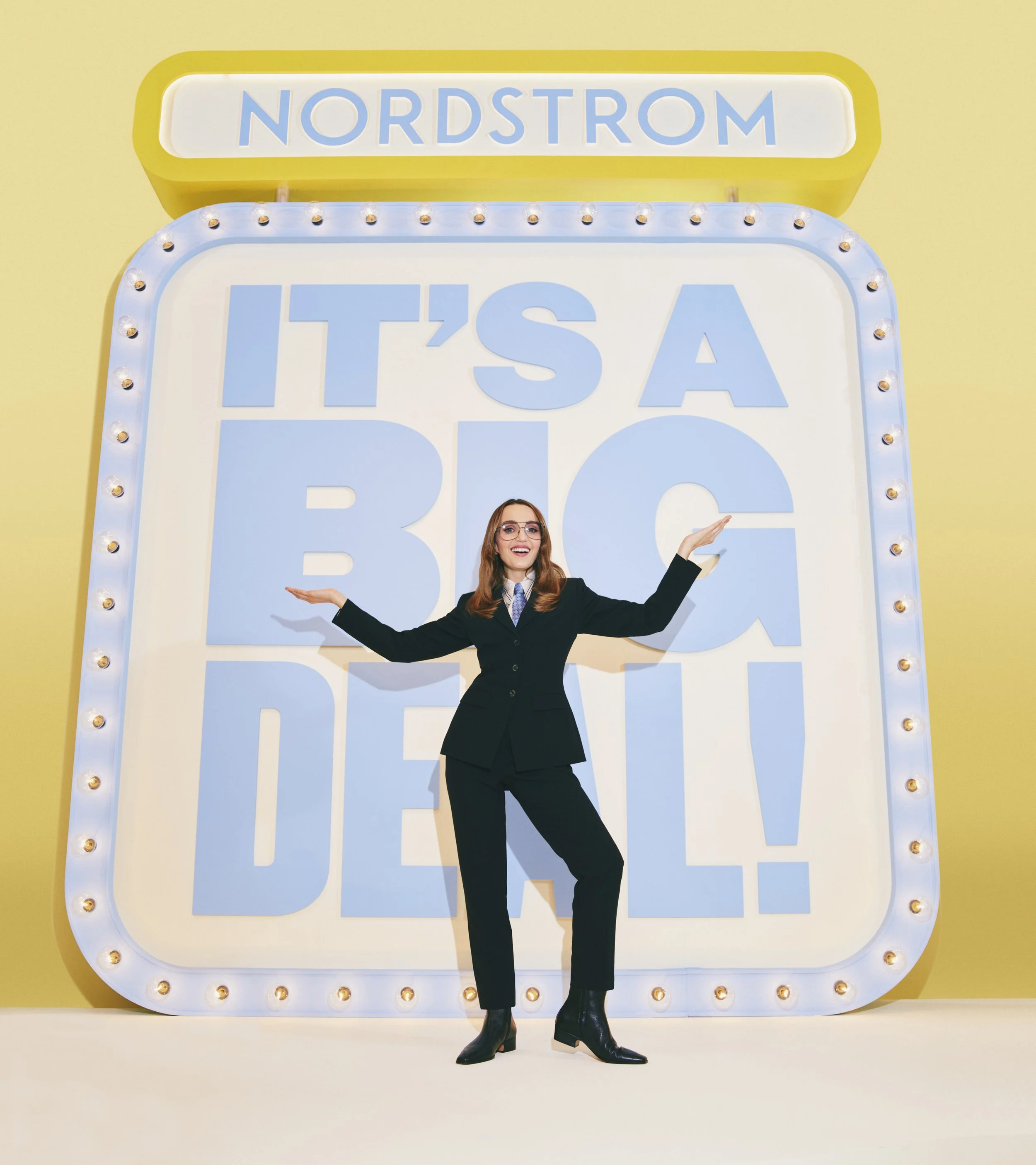 A woman with glasses and long red hair, dressed in a black blazer, white shirt, and black pants, standing in front of a large "Nordstrom" sign with lights around the border. She has her arms open wide and a big smile.