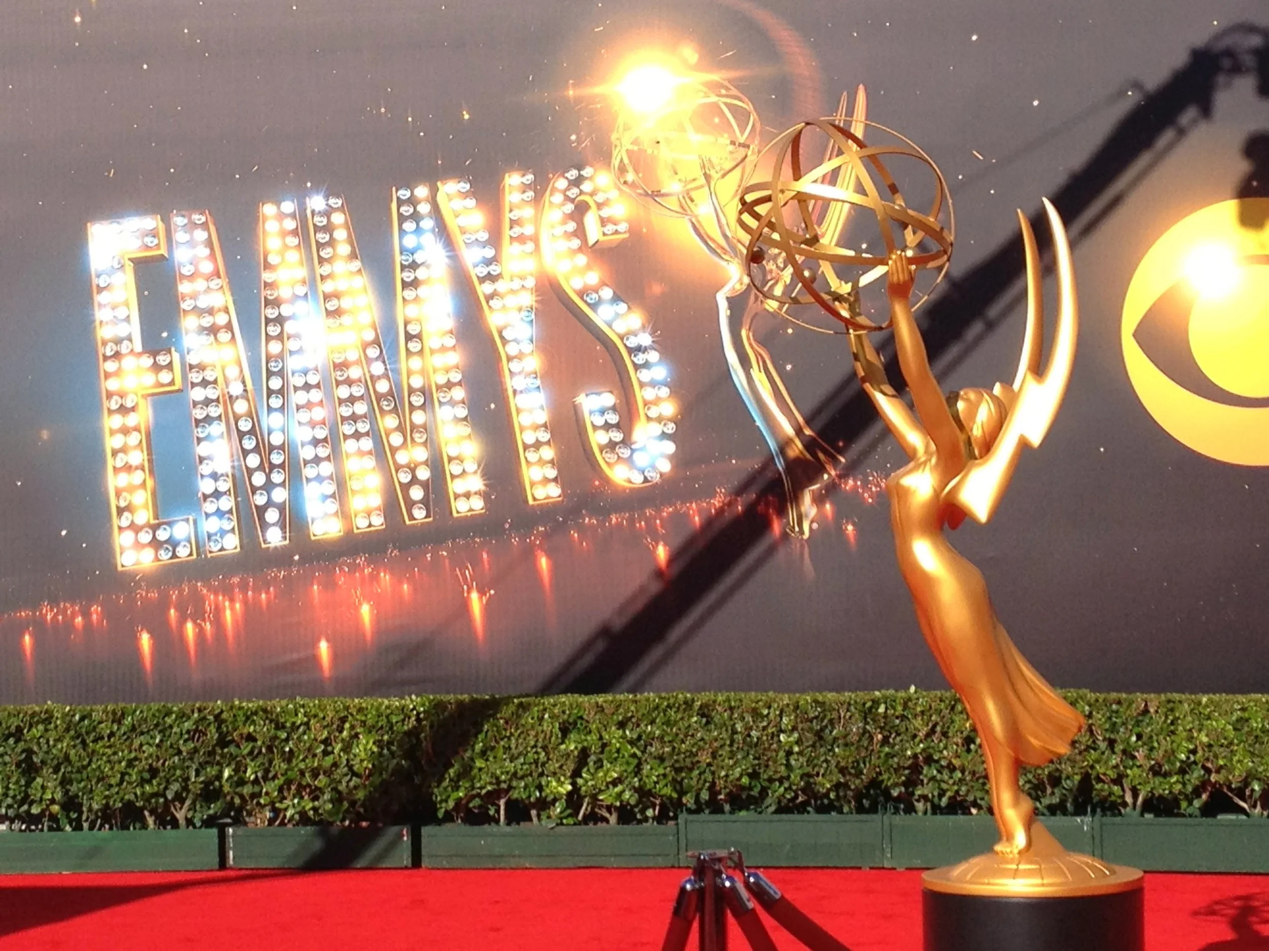 Golden Emmy Award trophy on red carpet with illuminated sign in background reading 'EMMY' with lights and sparkles.
