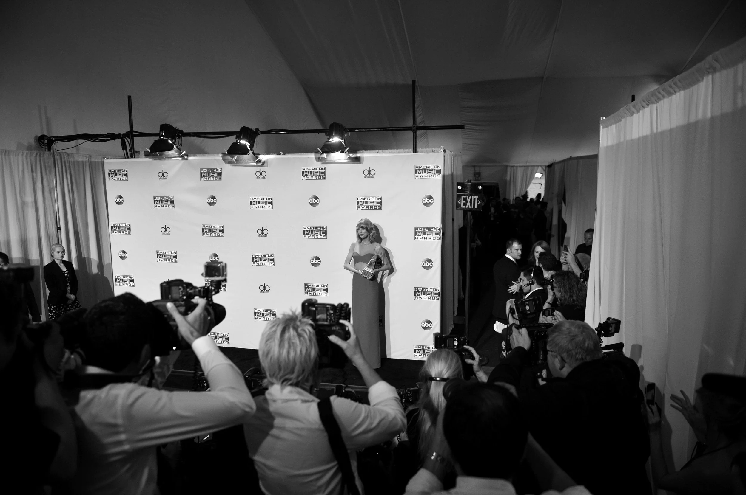 Black and white photo of a celebrity on stage at an American Music Awards event, with a backdrop displaying the event's logo and ABC. Audience members and photographers are visible in the foreground, capturing the moment.