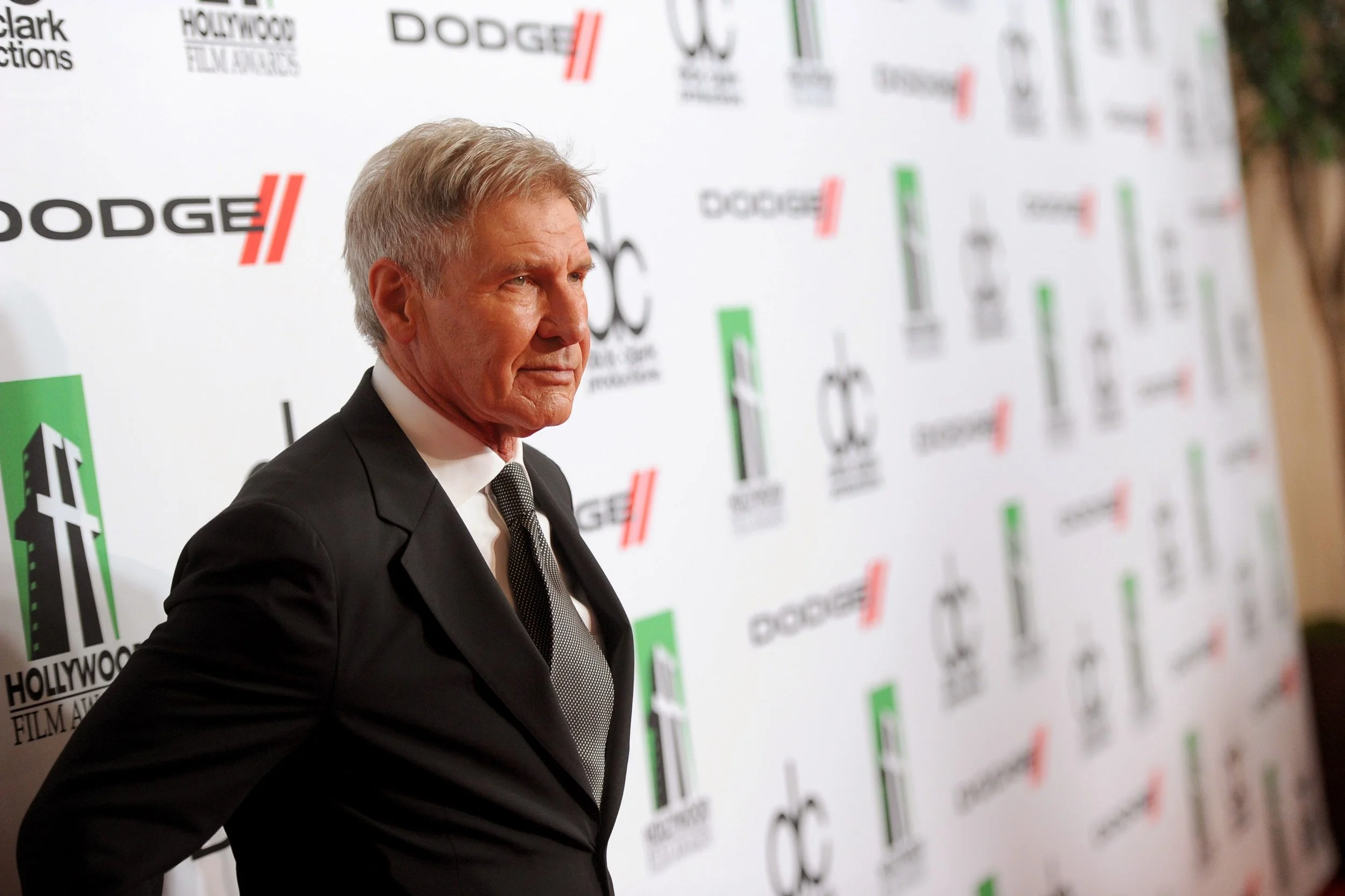 A man in a black suit and tie standing in front of a step-and-repeat banner with logos, including Hollywood, Dodge, and other sponsors, at a formal event.