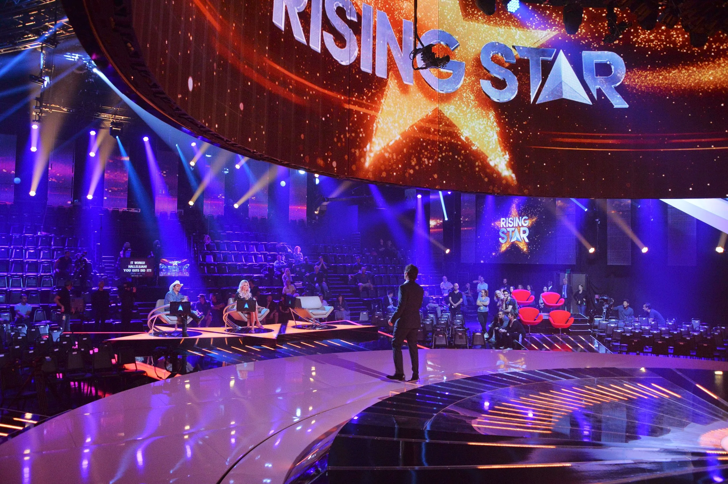 An indoor event space with a large curved LED screen displaying the words 'RISING STAR' surrounded by a star graphic and orange glowing effects. The stage features seated hosts or judges, with some audience members scattered throughout. Purple and bl