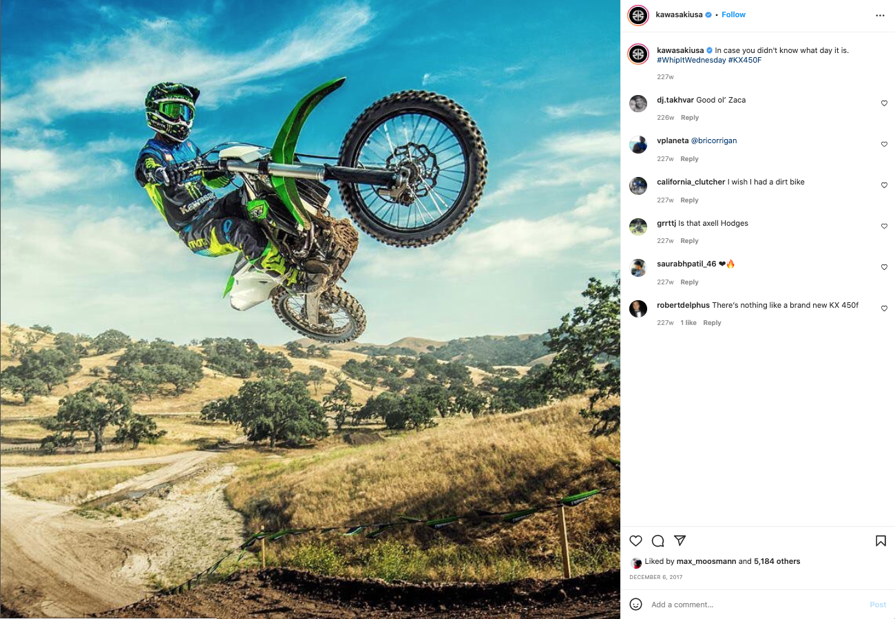 A motocross rider in blue and black gear performing a mid-air stunt on a green dirt bike against a background of rolling hills and trees under a partly cloudy sky.