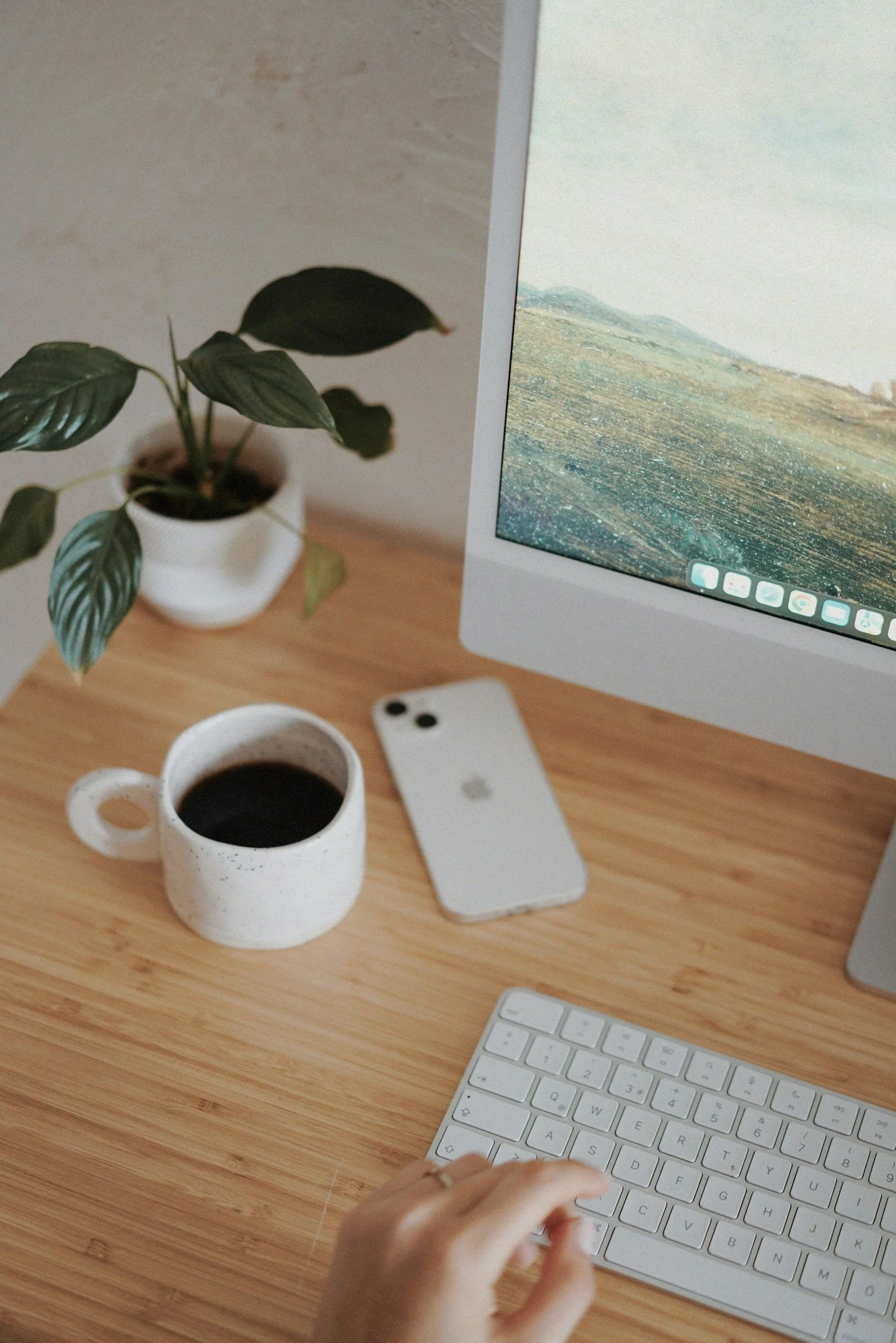 A person typing on a white keyboard with a wooden desk, a potted plant, two cups of coffee, a white iPhone, and a computer monitor displaying a landscape.