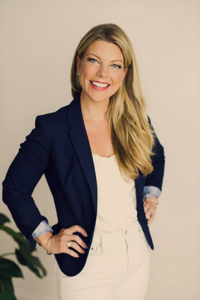 Jackie Phichith. A smiling woman with long blonde hair, wearing a navy blazer over a white top and white pants, standing with her hands on her hips against a plain background.