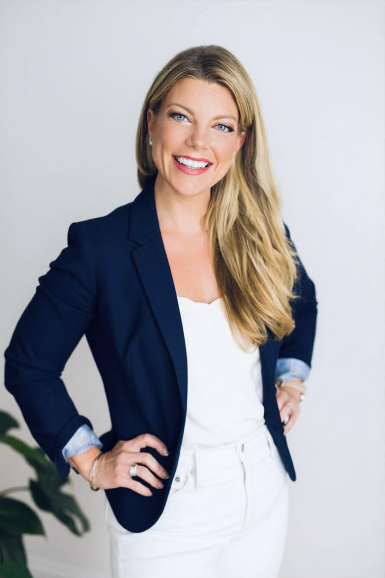 Jackie Phichith. A woman with long blonde hair, wearing a navy blazer over a white top and white pants, standing with her hands on her hips, smiling against a plain white background.