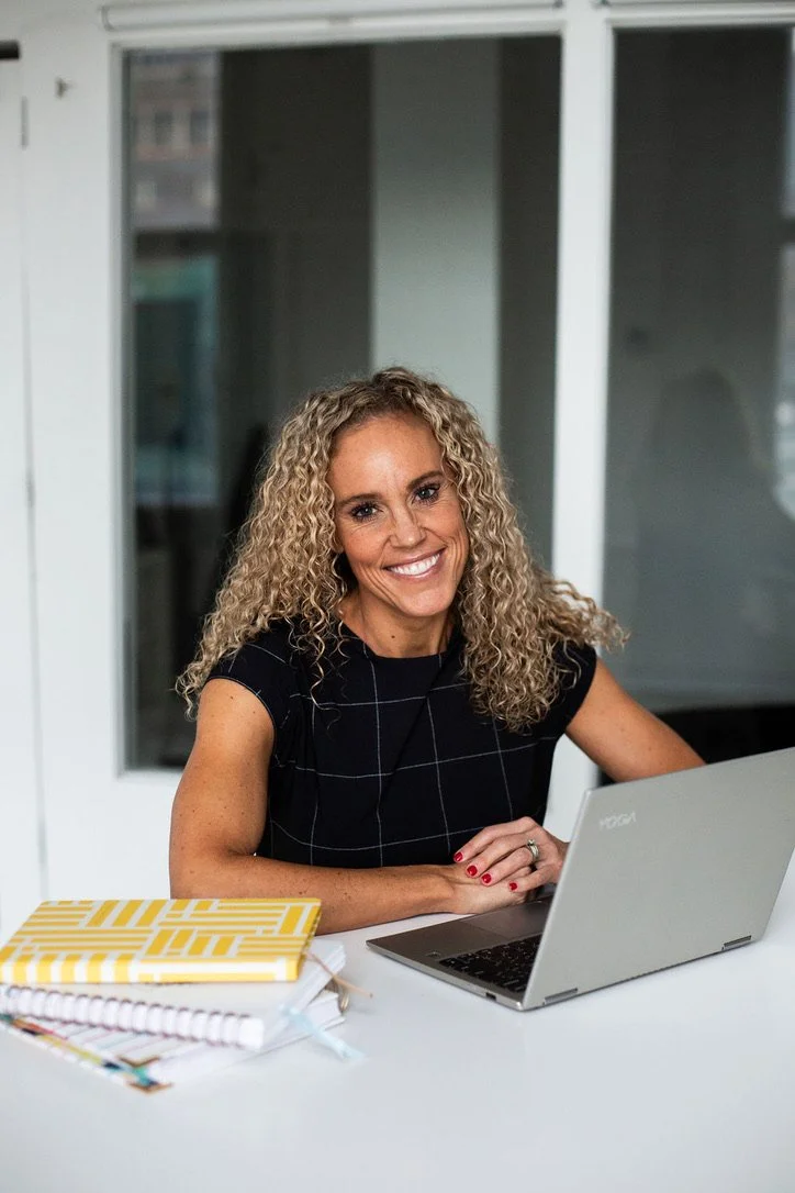 A smiling woman with curly blonde hair working on a silver laptop at a desk with notebooks and a face mask.