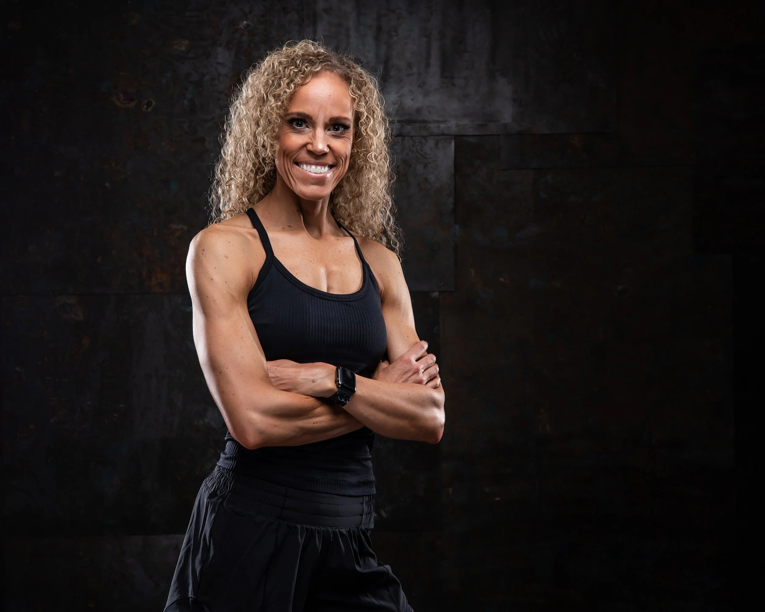 A fit woman with curly blonde hair standing against a dark backdrop, wearing a black sports top and black athletic pants, smiling with arms crossed.