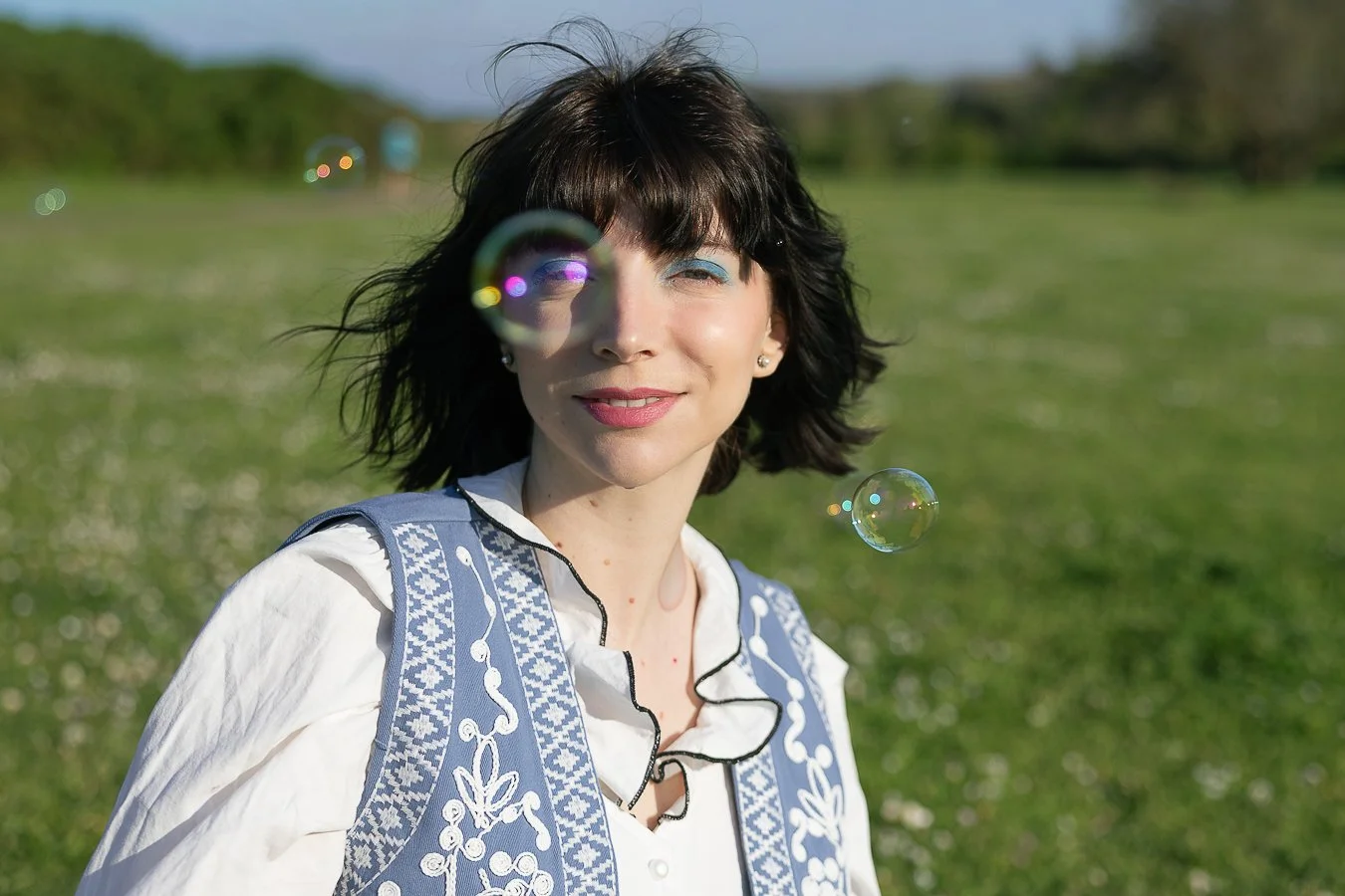 Una donna con capelli castani corti e vestito bianco con ricami blu, che sorride all'aperto in un campo verde con bolle di sapone.