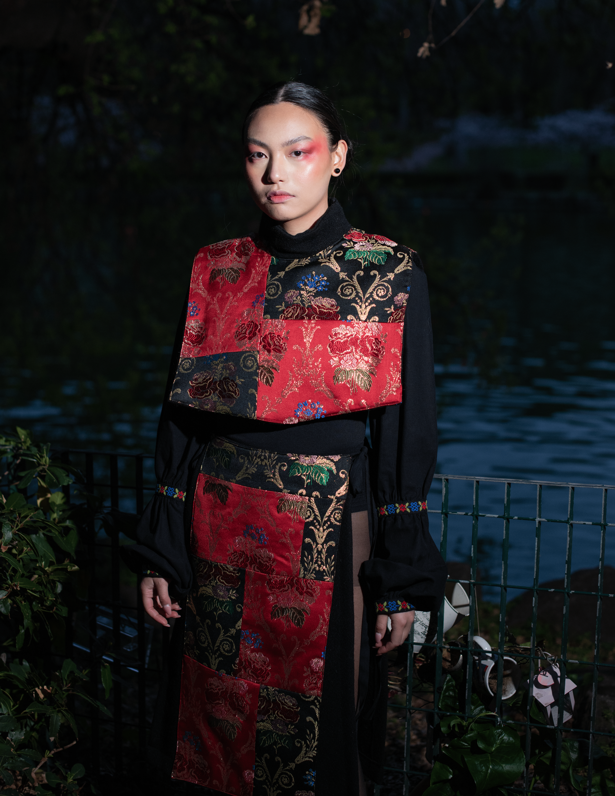 Una donna con trucco artistico sui occhi indossa un vestito con dettagli ricamati di vari colori, sullo sfondo di un'area verde e acqua in condizioni di scarsa illuminazione.