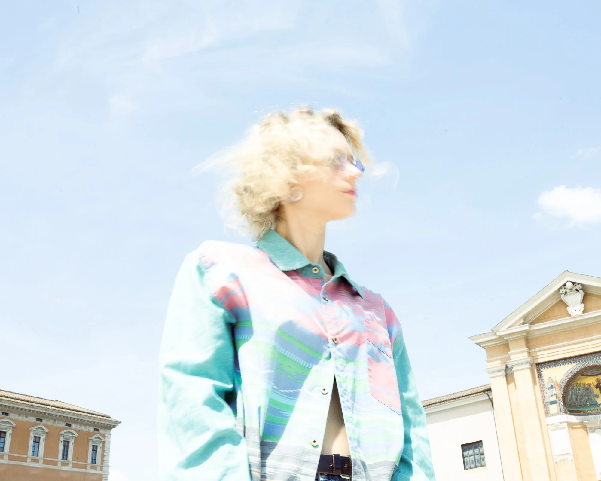 Ragazzo con capelli biondi e riccioli corti, indossa una camicia colorata con motivi a righe di diversi colori. Sullo sfondo, un edificio storico con archi decorativi e dettagli ornamentali, sotto un cielo senza nuvole.