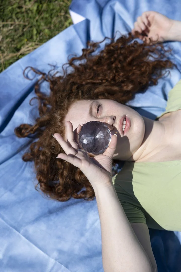 Una ragazza con capelli ricci rossi sdraiata su un plaid blu in mezzo all'erba, che tiene una grande sfera di cristallo davanti all'occhio.
