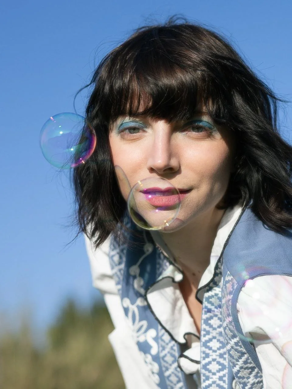 Una donna con capelli castani corti, indossa trucco azzurro e una camicetta bianca con dettagli azzurri, sorride verso la fotocamera mentre ci sono bolle di sapone nell'aria, con uno sfondo di cielo blu.