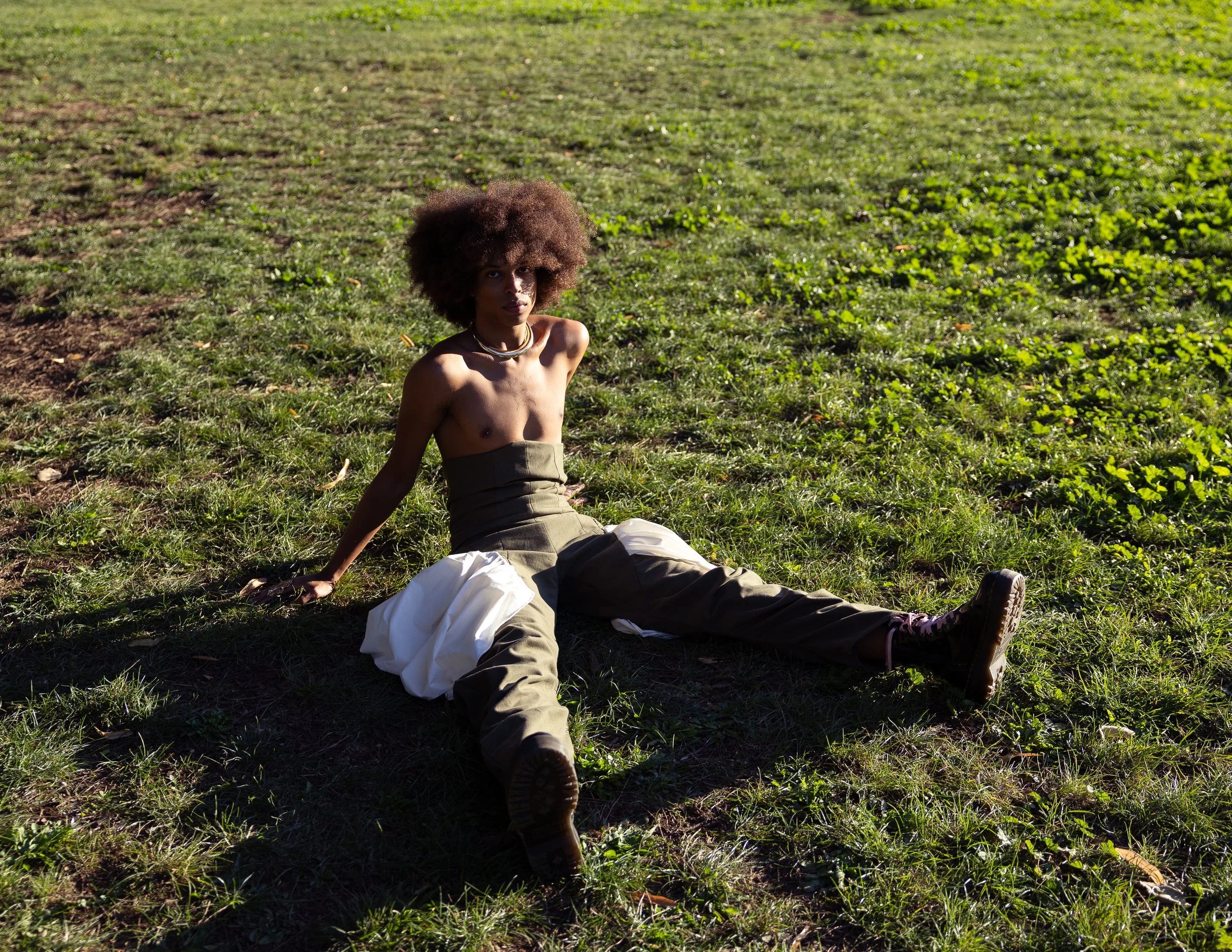 Ragazza con capelli afro seduta sull'erba in un campo, indossa pantaloni cargo e una collana, con uno sfondo di vegetazione verde e terra.