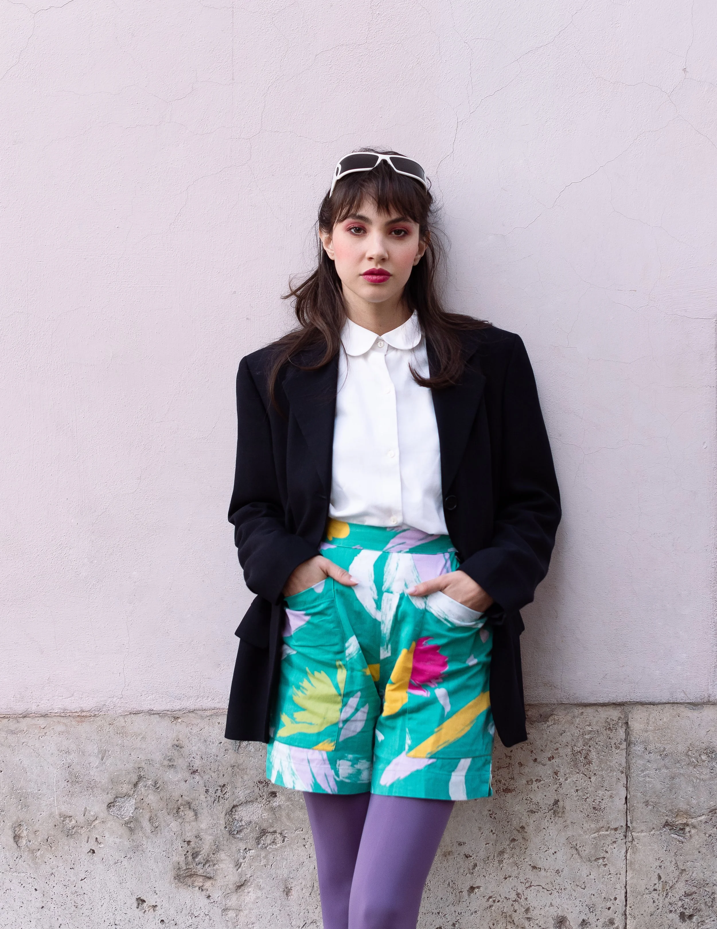 Una donna con capelli castani corti e trucco rosa, indossa occhiali da sole sulla testa, una camicetta bianca, una giacca nera, una gonna colorata e pantaloni viola, si appoggia a un muro con scritto crack.