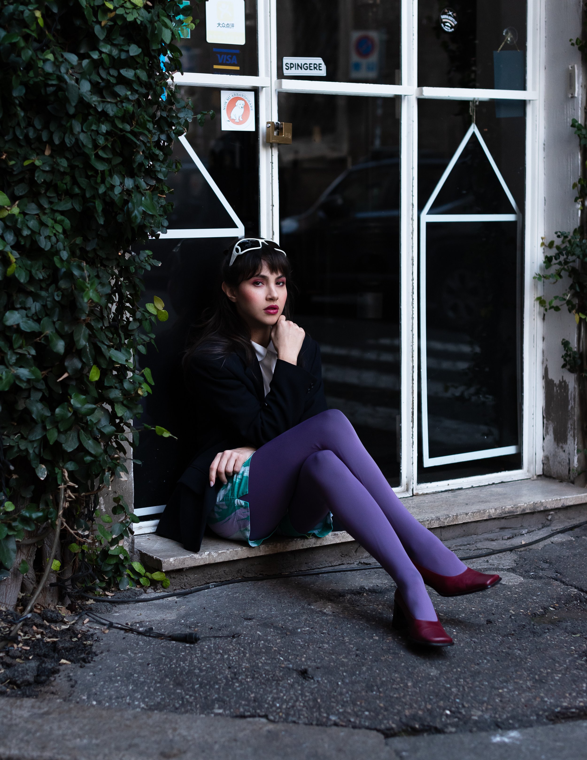 Una donna seduta su uno scalino davanti a una porta di vetro, con folti capelli castani e occhiali sulla testa, indossa una giacca nera, una minigonna a quadri, calze viola e scarpe bordeaux. Accanto a lei ci sono piante verdi e un pavimento in pietr