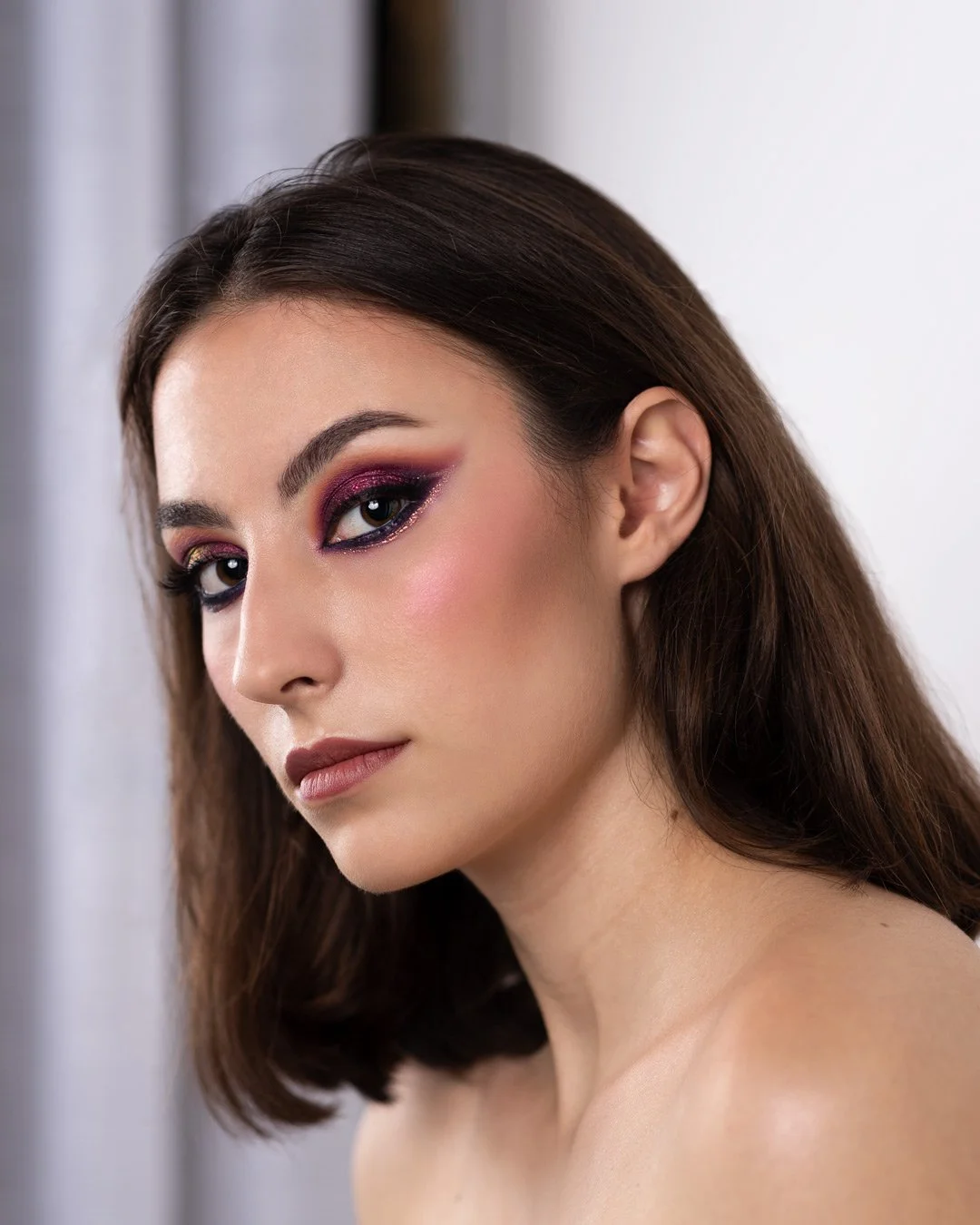Ragazza con trucco occhi glitterato rosa e viola, capelli castani mossi, pelle chiara, sfondo neutro.