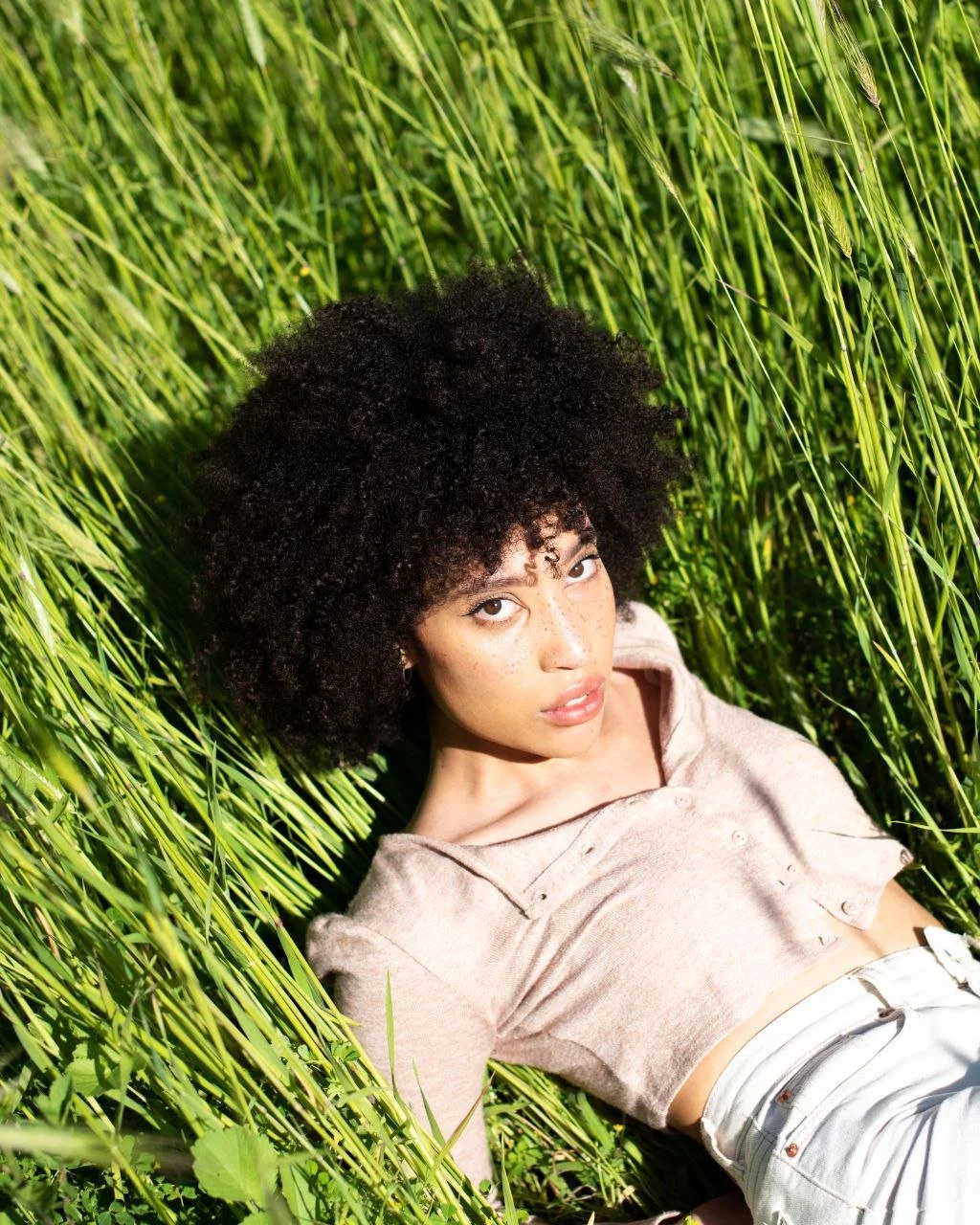 Ragazza con capelli ricci neri e viso con lentiggini giace in un campo di grano verde.