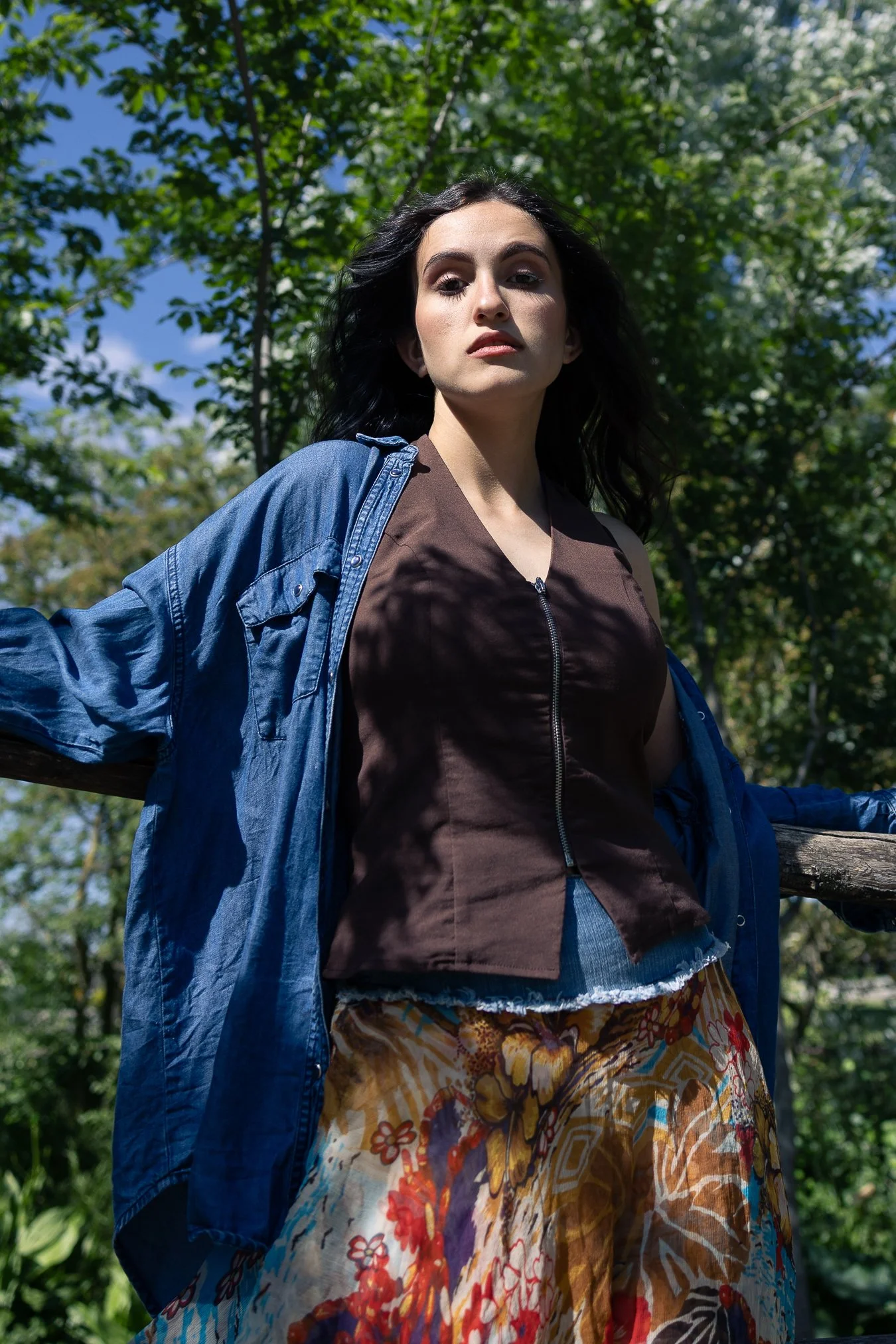 Ragazza con capelli neri, vestito marrone con zip, giacca blu, gonna colorata con motivi floreali e sfondo di alberi e cielo.