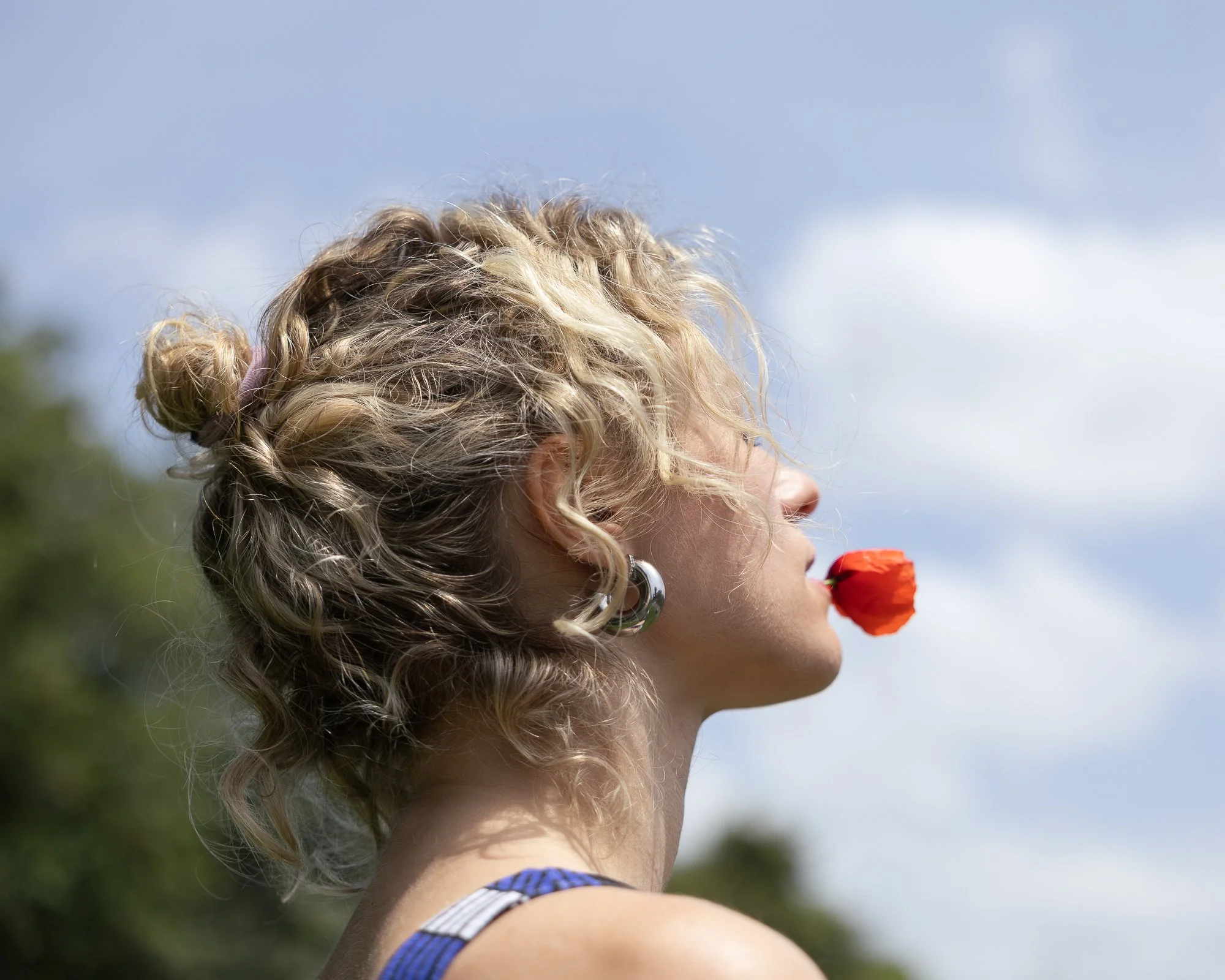 Prima piano di una donna con capelli ricci biondi che tiene un fiore rosso nella bocca, vista laterale, sotto un cielo chiaro.
