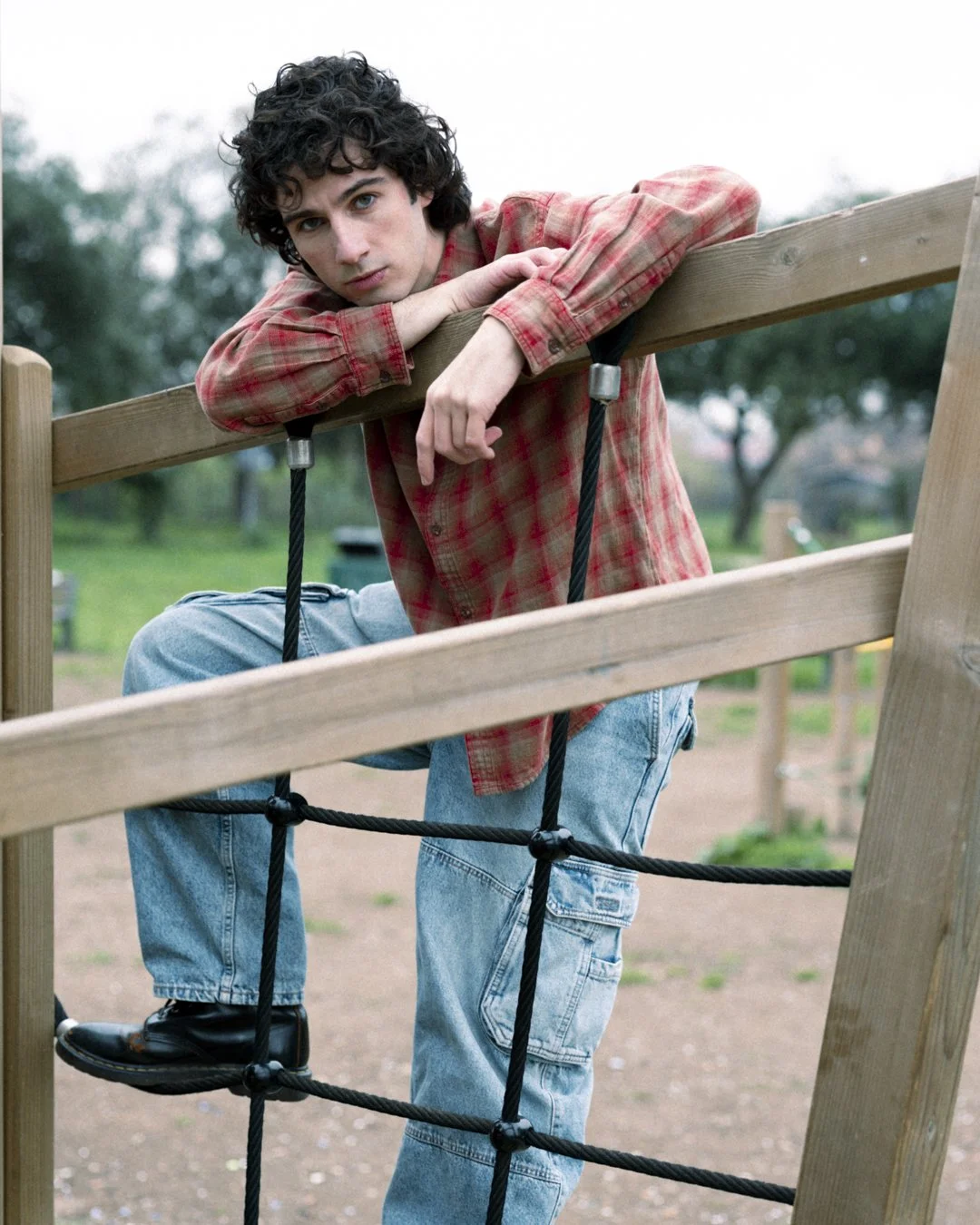 Ragazzo con capelli ricci, camicia a quadri rossa e jeans semplici, appoggiato a una struttura da parco giochi in legno e corda, con uno sfondo verde di alberi e percorso in terra martoriata.