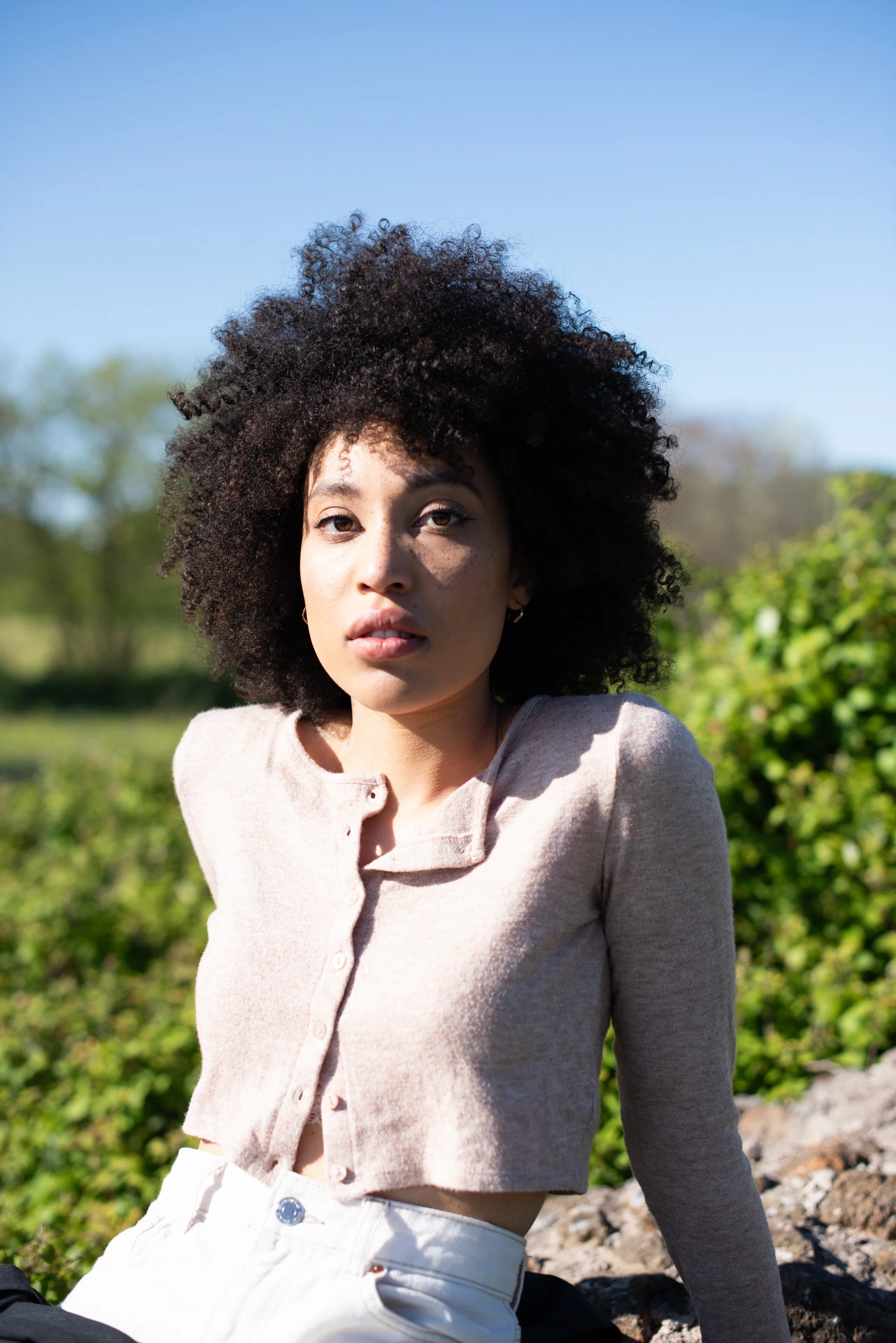 Ragazza con capelli ricciari e pelle chiara, indossa una maglietta beige, si trova all'aperto con cielo blu e vegetazione verde sullo sfondo.