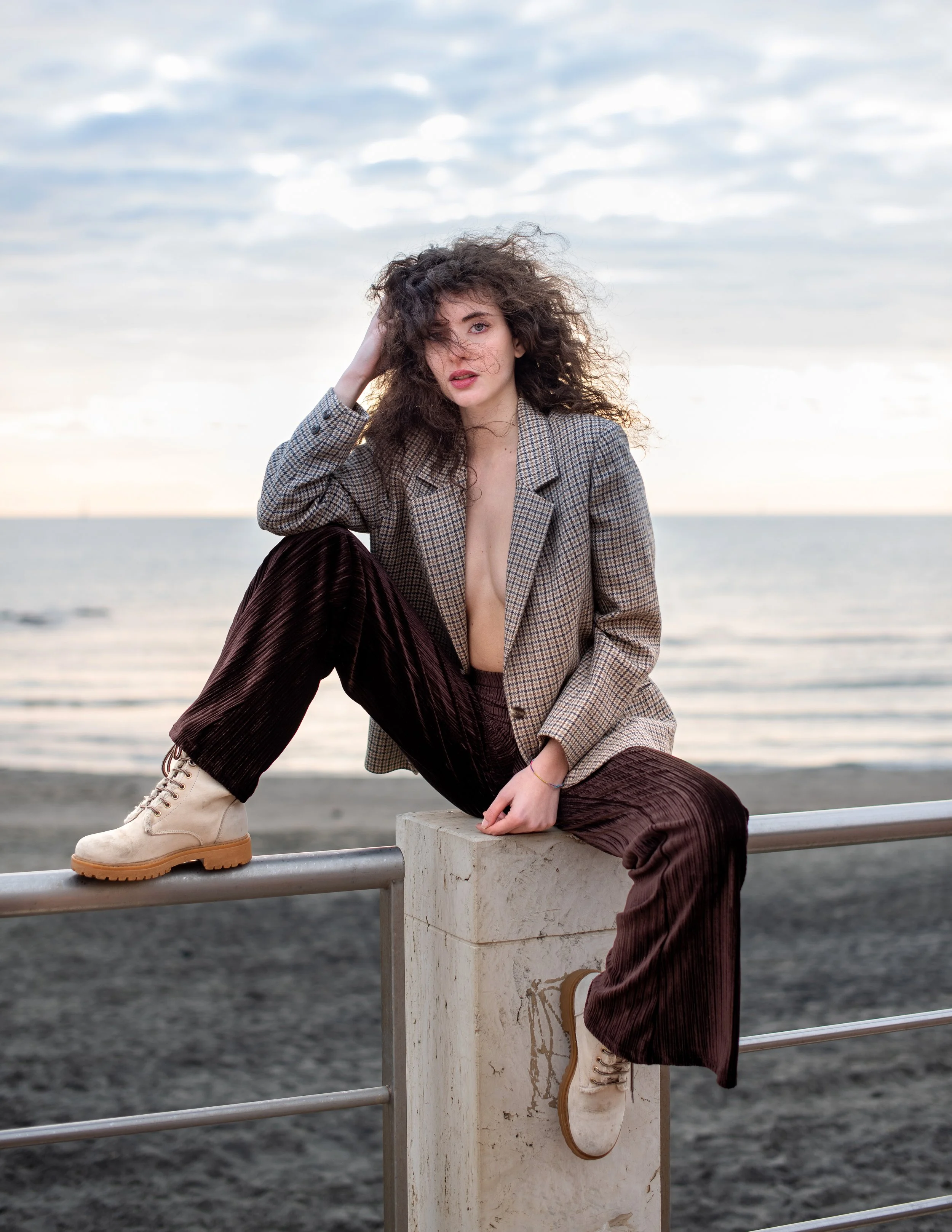 Una donna seduta su un block di cemento vicino al mare con le mani tra i capelli, indossa un blazer a quadri, pantaloni a righe e scarpe chukka beige, con uno sfondo di nuvole e mare.