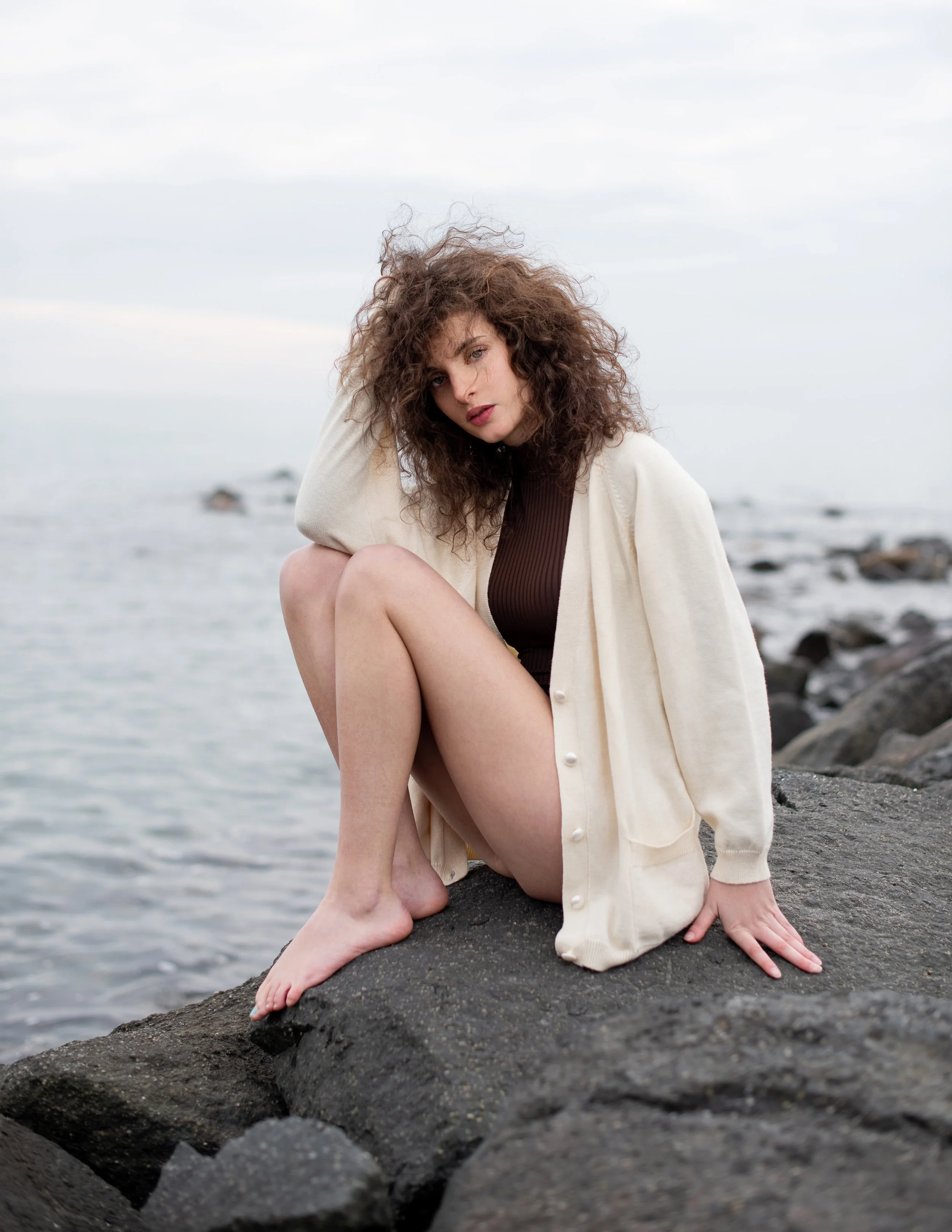 Donna con capelli ricci e mossi, seduta su una roccia sulla spiaggia, indossa una maglietta marrone e un cardigan beige. Sullo sfondo si vede il mare con alcune rocce.