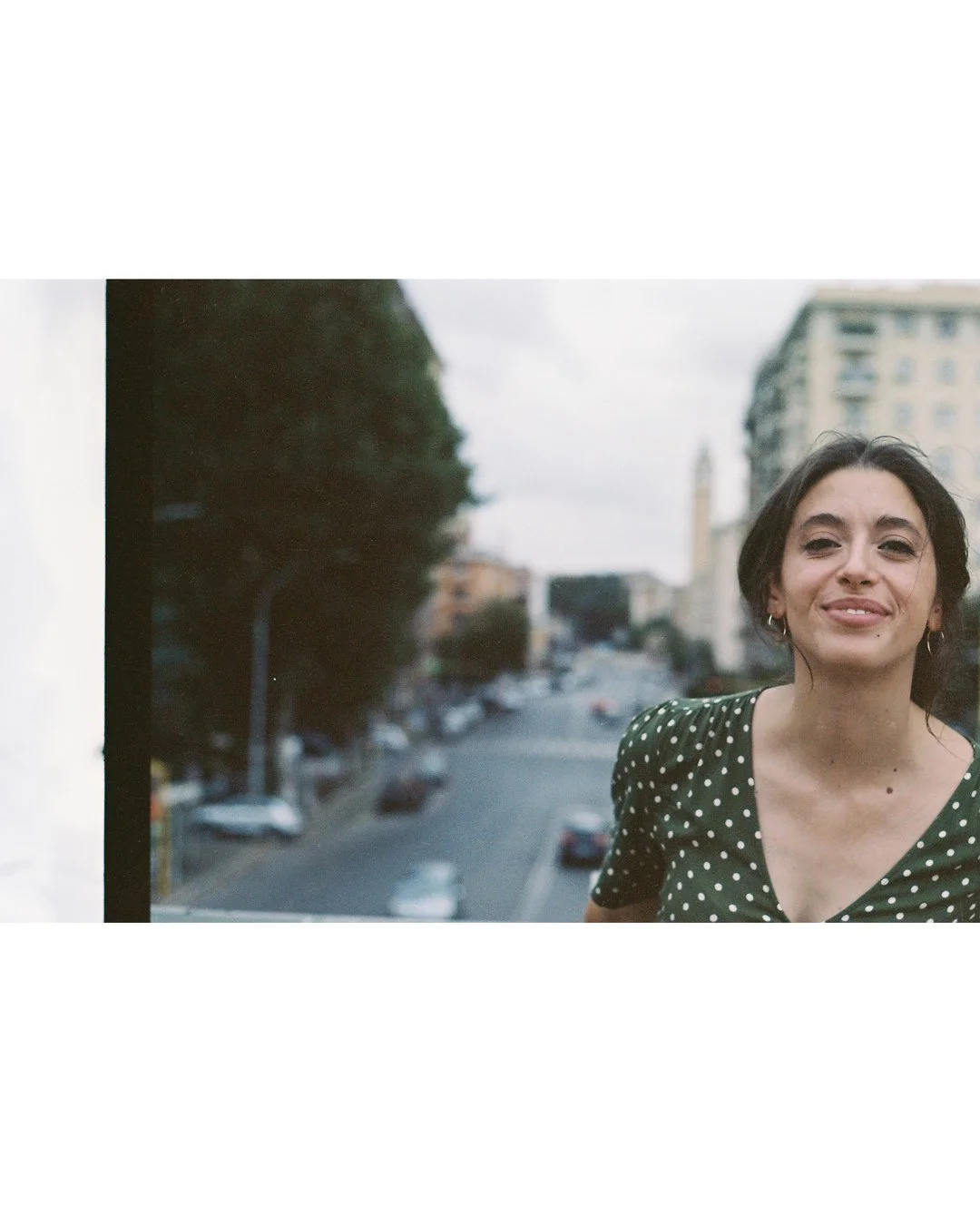 Una donna sorridente con capelli scuri corti, indossa una maglietta verde con pois bianchi, si trova davanti a una finestra con vista sulla città