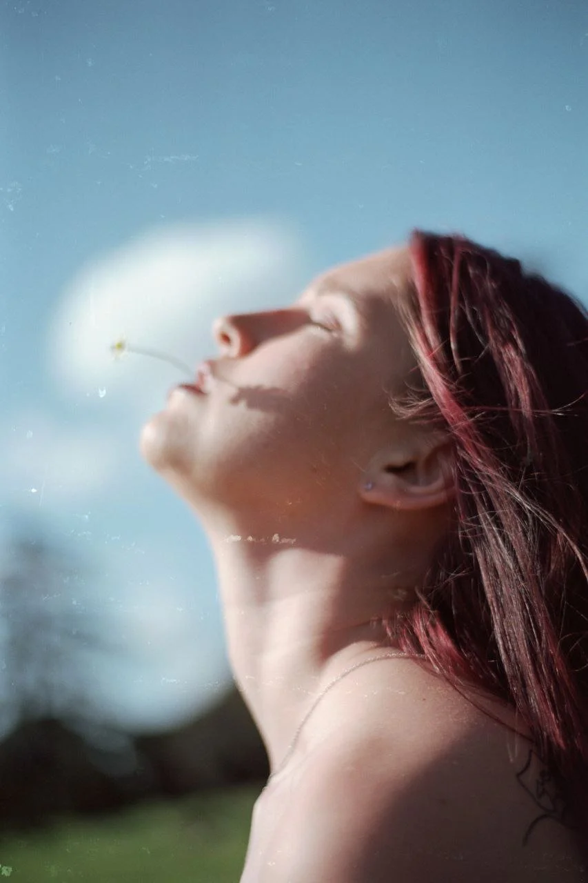 Una ragazza con i capelli rossi e abbronzata, con gli occhi chiusi, tiene una margherita vicino al viso sotto un cielo blu.