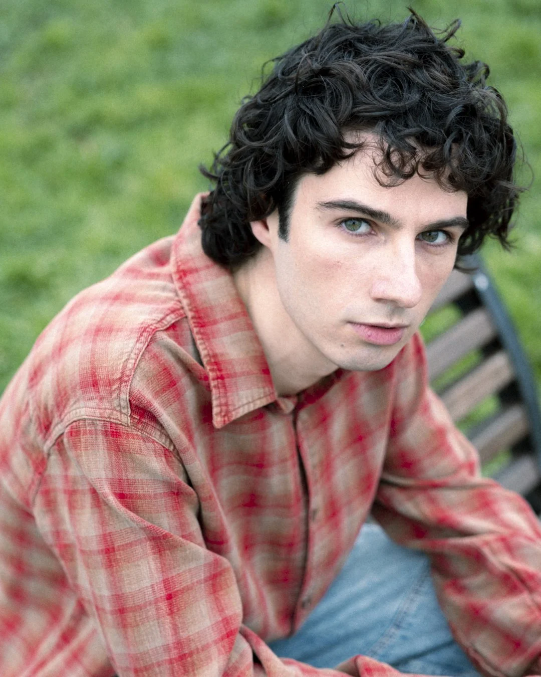 Ragazzo con capelli ricci e occhi azzurri seduto su una panchina in un prato verde, indossa una camicia a quadri rossa e arancione.