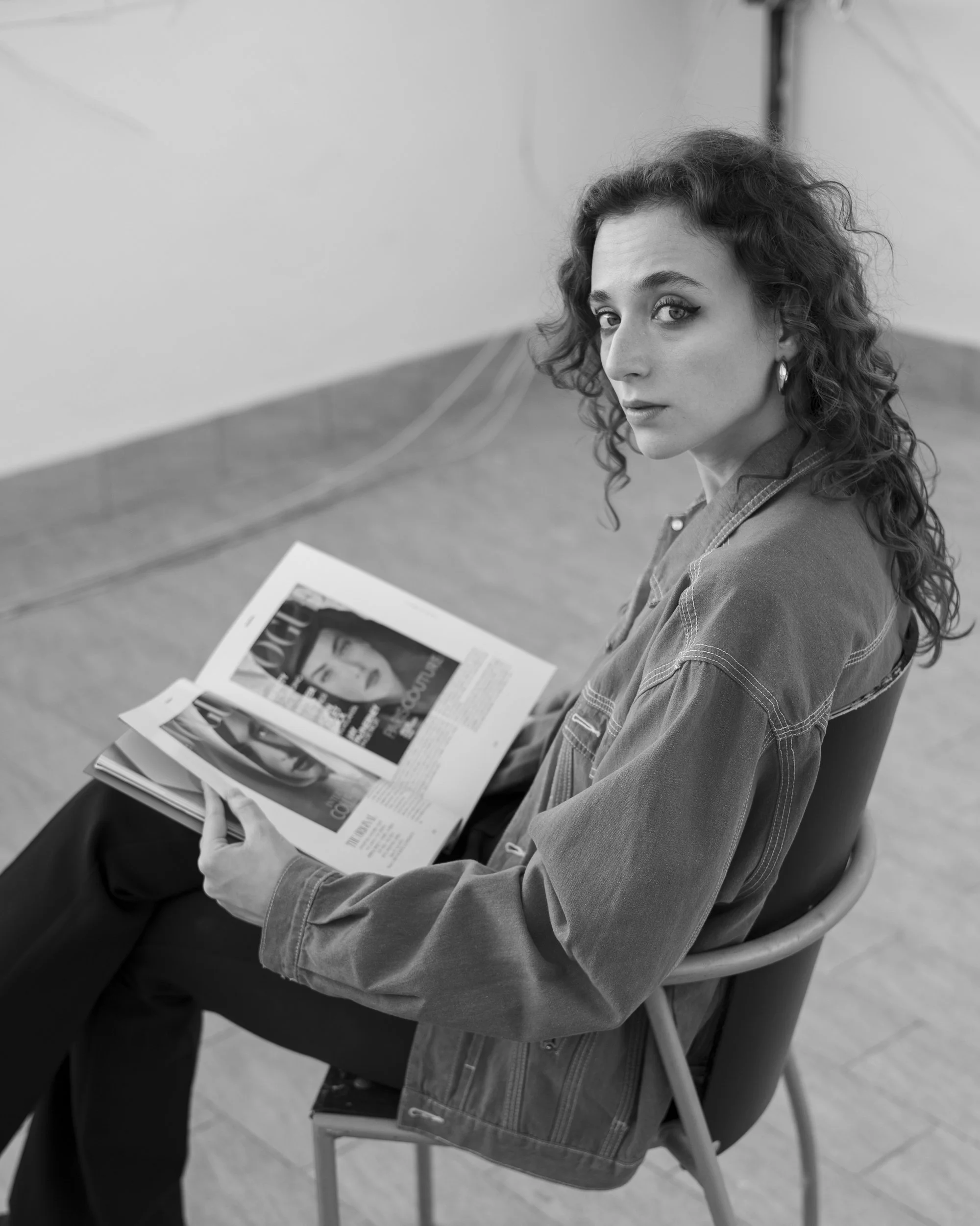Una donna seduta su una sedia di plastica, che guarda verso l'obiettivo mentre tiene un giornale aperto sulle ginocchia.