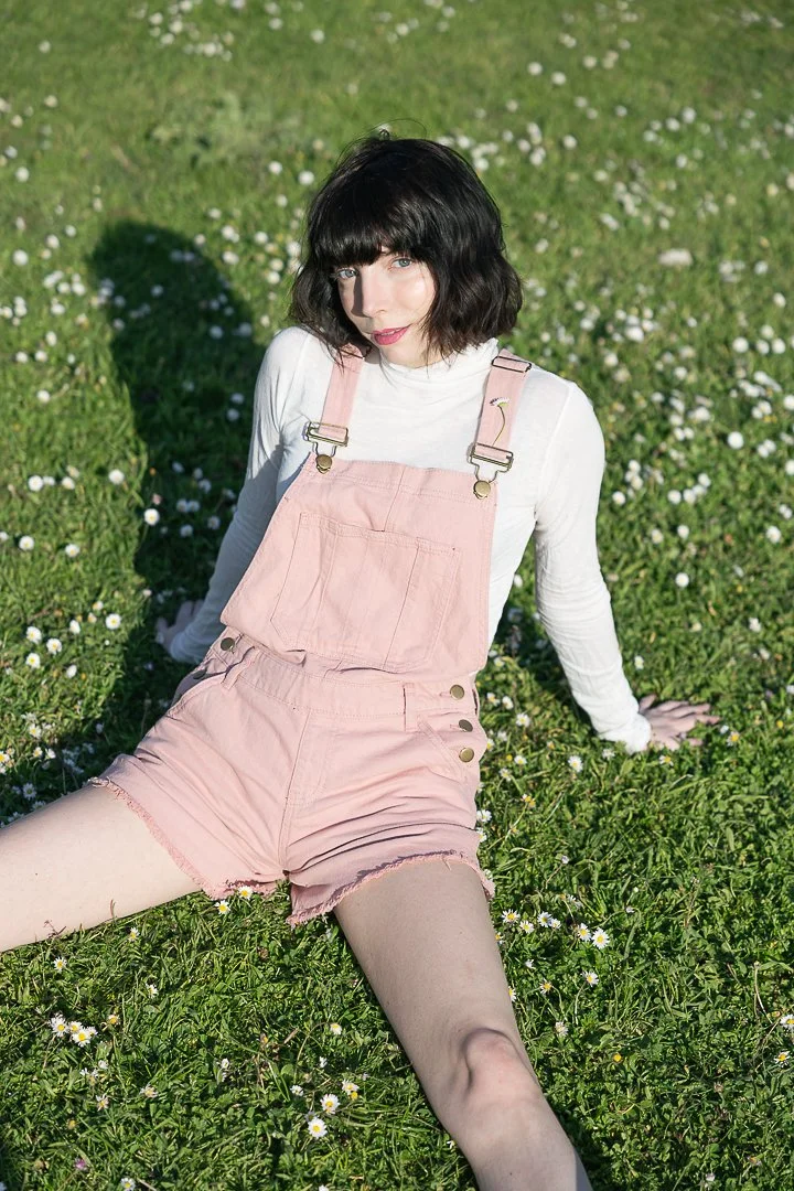 Ragazza seduta sull'erba con fiori bianchi, indossa un maglione bianco e salopette rosa, ci sono alberi e cielo sullo sfondo.