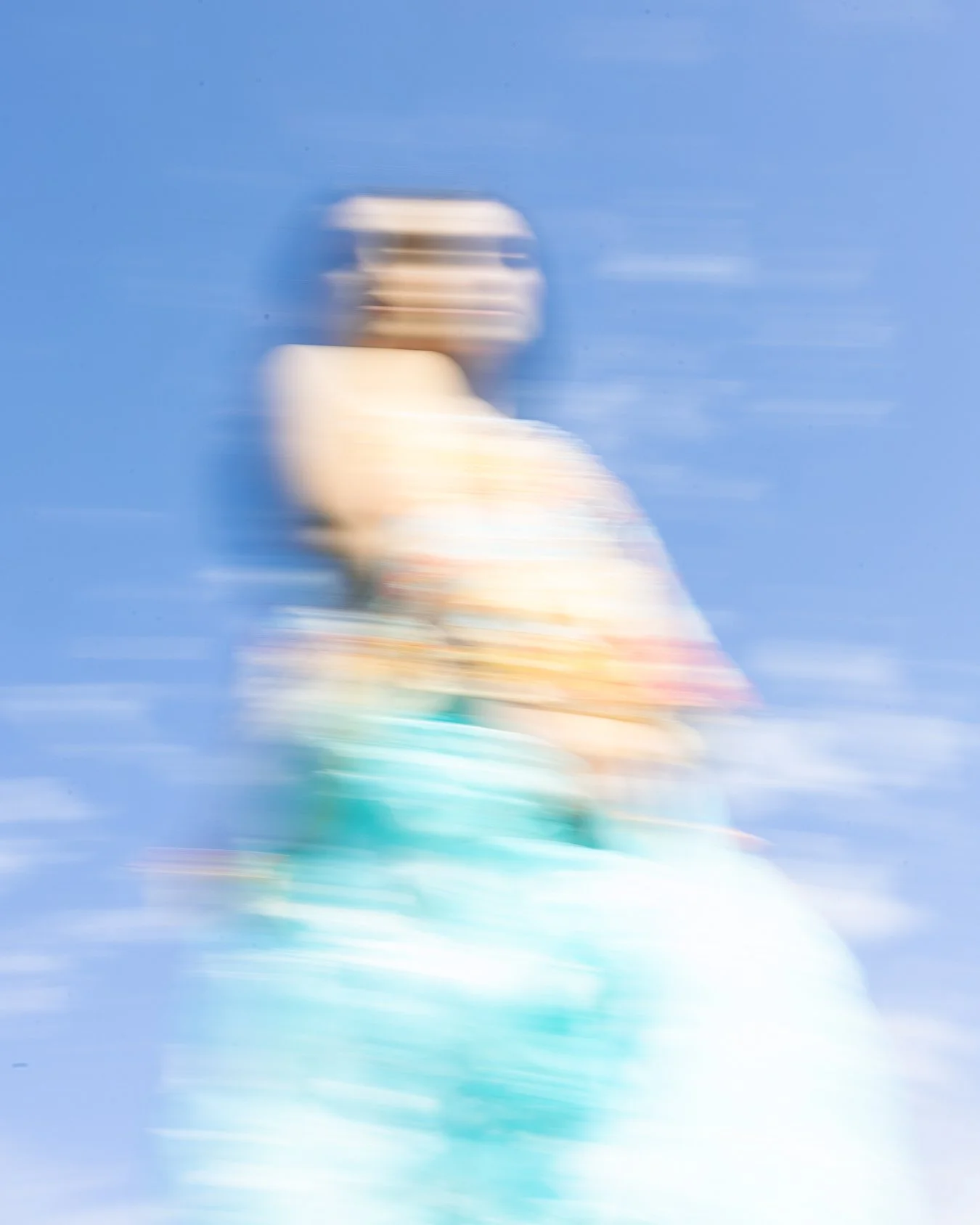 Immagine sfocata di una donna con capelli corti, vestita con abiti colorati, che cammina all'aperto contro un cielo blu, con movimento rapido.