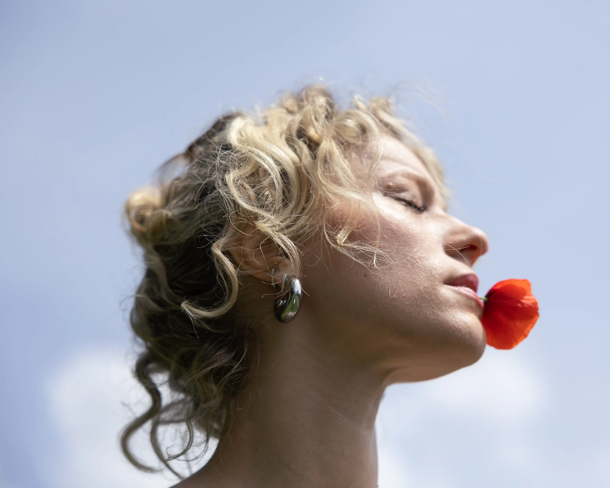 Profilo di una donna con occhi chiusi, capelli ricci biondi, che tiene un fiore rosso vicino al viso, sfondo di cielo blu.