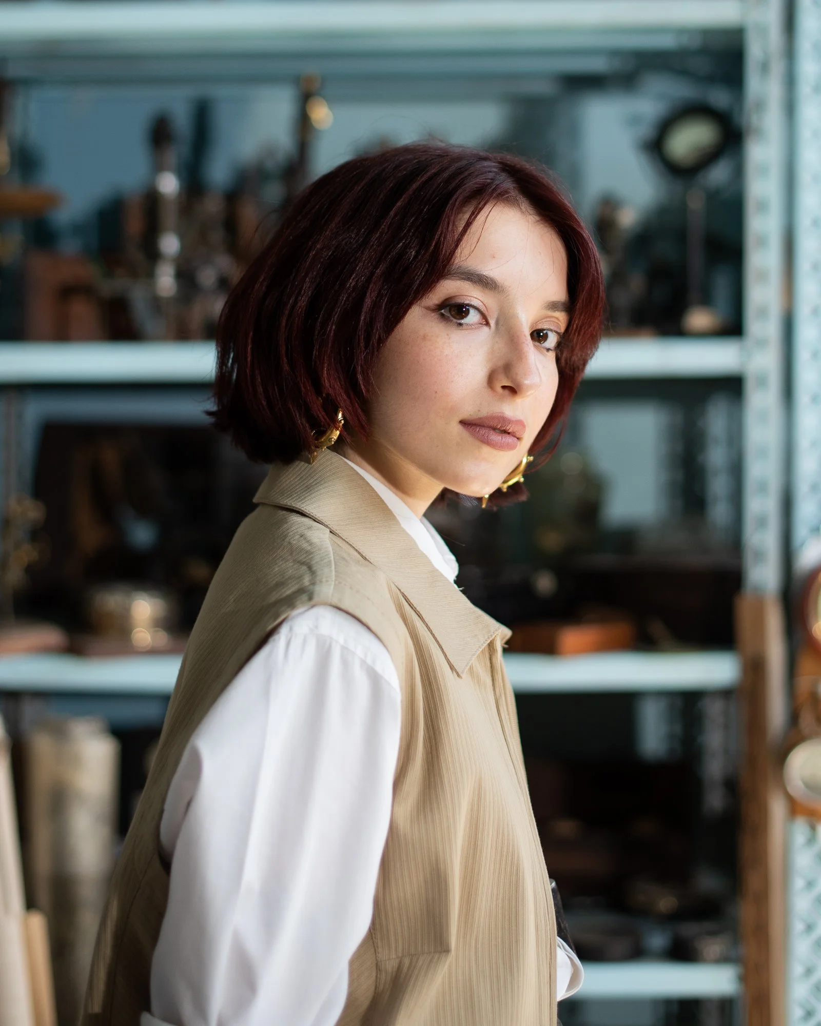 Una donna con i capelli castani corti e occhi marroni guarda verso l'obiettivo, indossa indumenti color beige e bianco, nell'ambiente di un laboratorio o studio con scaffali e strumenti sullo sfondo.