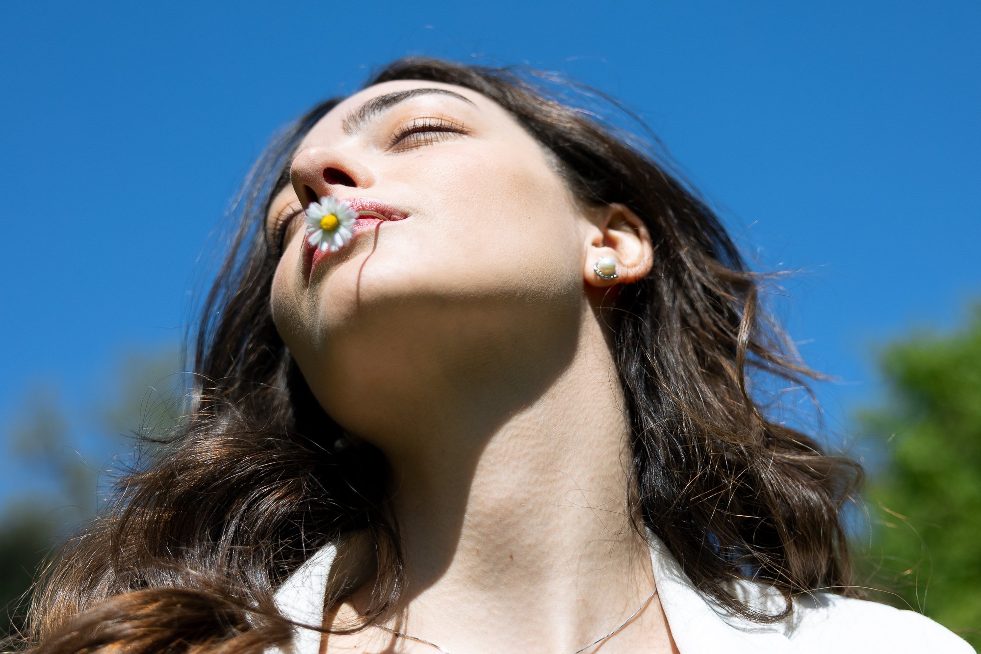 Ragazza con capelli ricci, occhi chiusi, che tiene un fiore sulla bocca e sorride leggermente, sfondo di cielo blu e qualche albero.