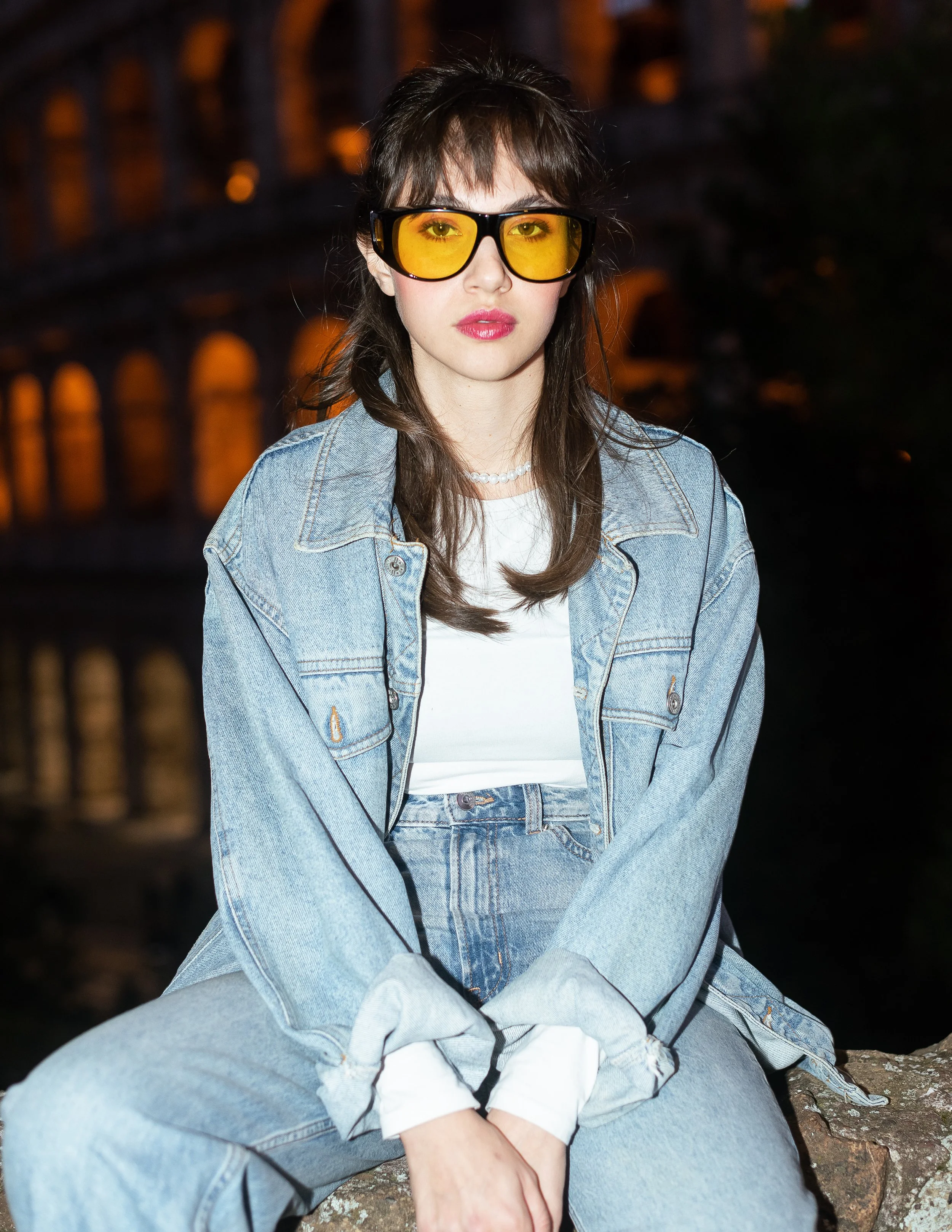 Ragazza con occhiali da sole gialli, maglietta bianca, giacca di jeans e jeans, seduta su una roccia di sera.