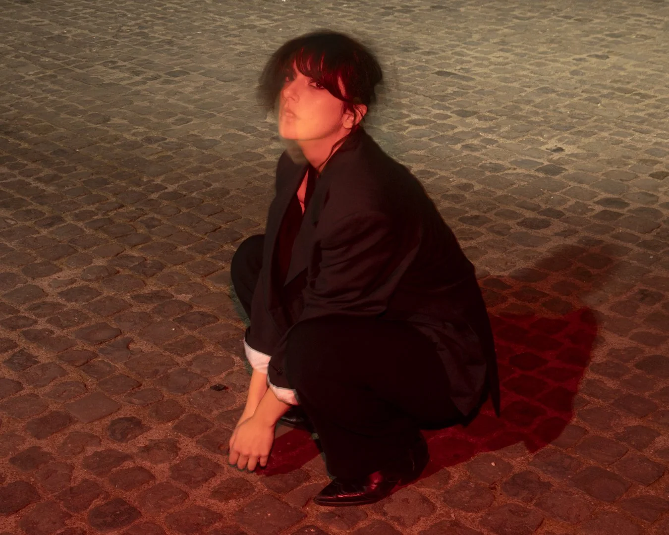 Una persona vestita di nero con capelli scuri e corti, seduta a terra su un marciapiede di pietra, con una luce rossa che illumina la scena.