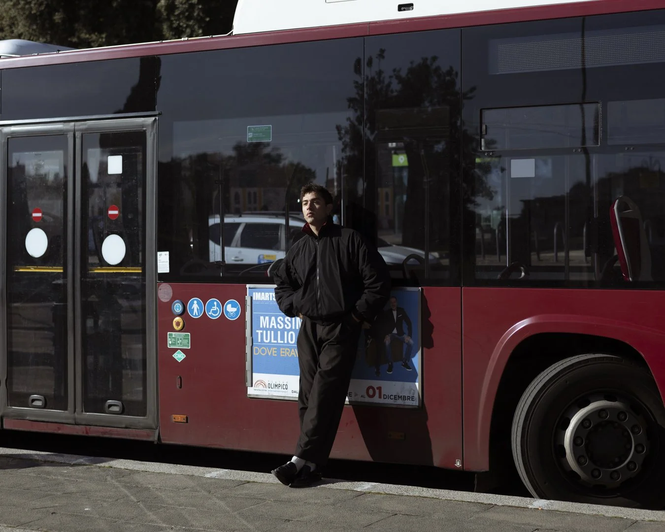 Un giovane uomo si appoggia a un autobus rosso in attesa, con uno sfondo di strada e alberi.