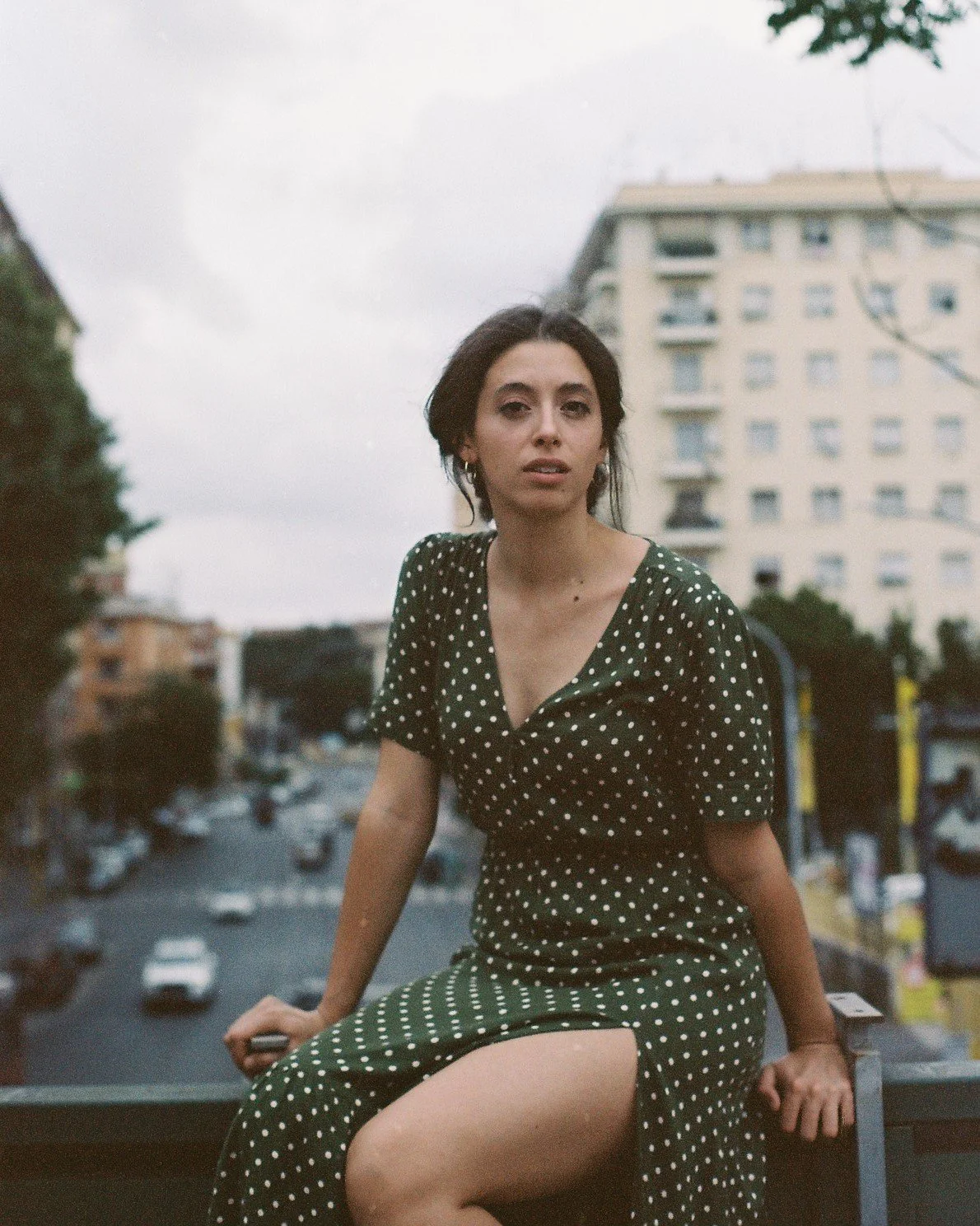 Una donna con un vestito verde con pois bianchi siede su una struttura metallica in una città, sullo sfondo si vedono alcuni edifici e un cielo nuvoloso.