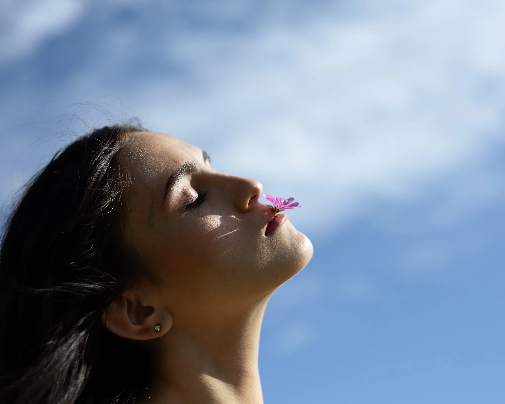 Profilo di una donna con gli occhi chiusi che tiene un fiore rosa vicino al volto, sotto un cielo azzurro con nuvole