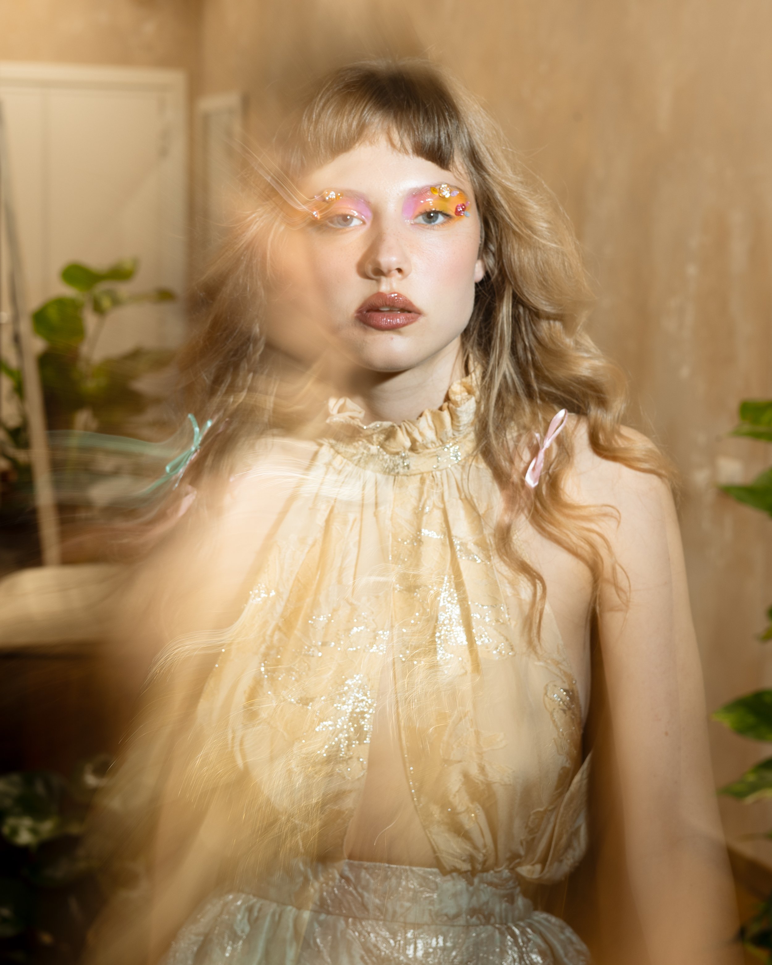 Ragazza con trucco colorato e decorazioni floreali sugli occhi, indossa un vestito chiaro su uno sfondo neutro, con alcune piante visibili ai lati.