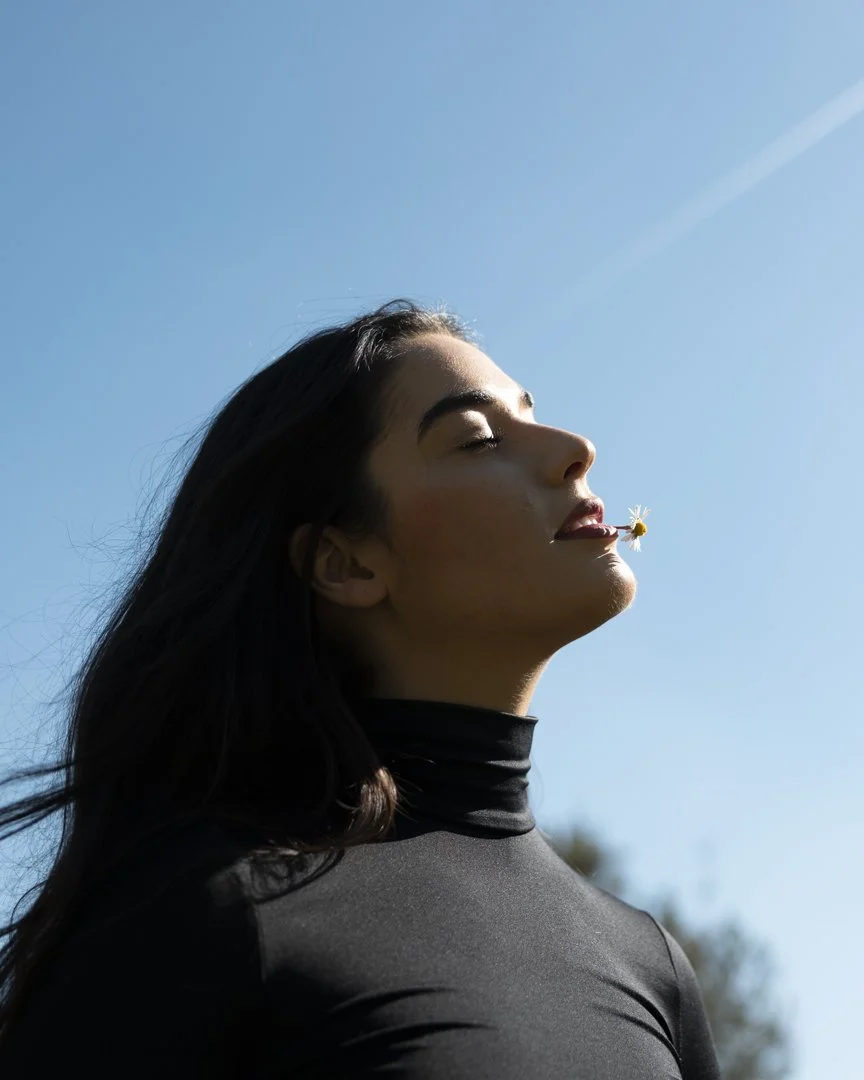 Ragazza con i occhi chiusi e bocca leggermente aperta, con un fiore tra le labbra, contro un cielo azzurro chiaro.