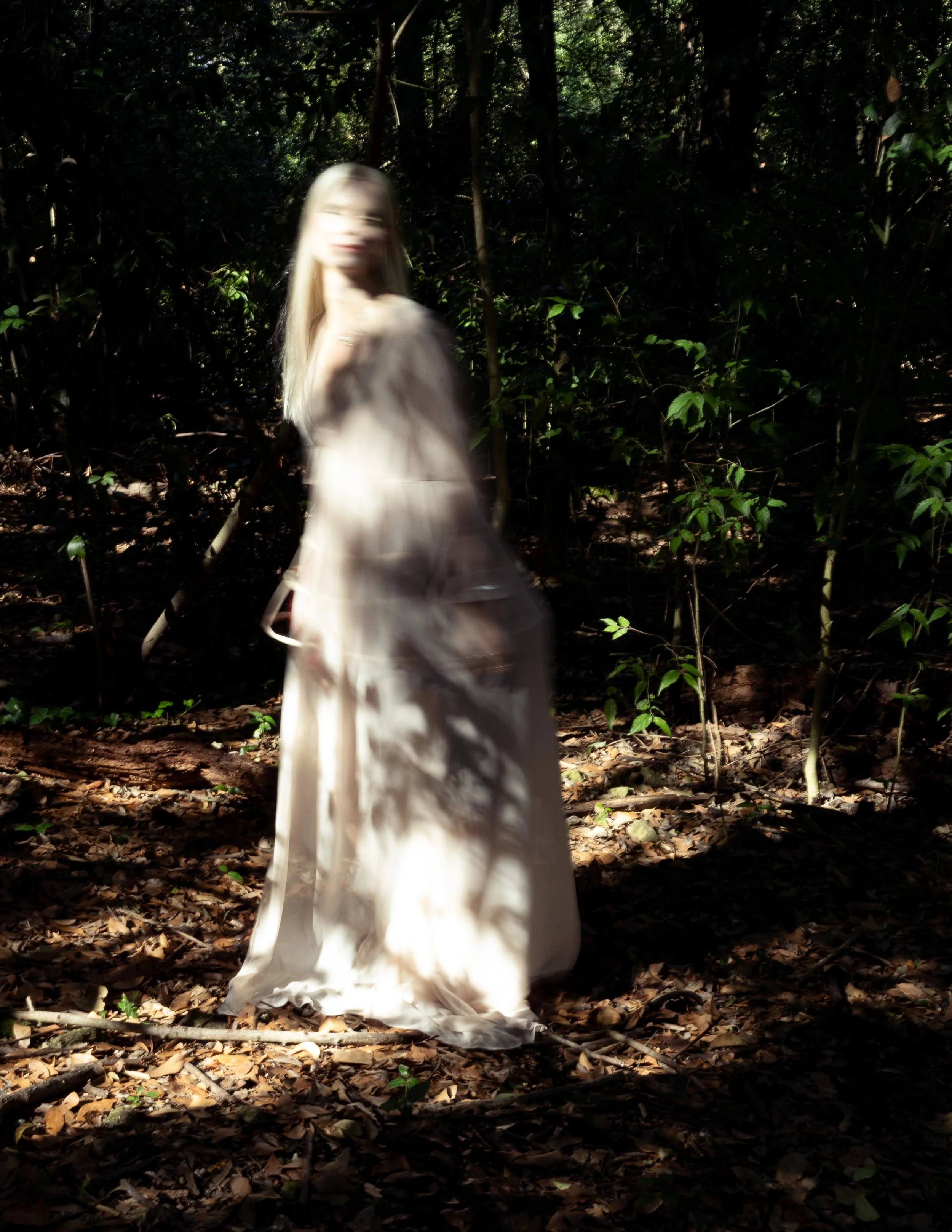 Figura femminile in abito bianco in un bosco, con luci e ombre che filtrano tra gli alberi.