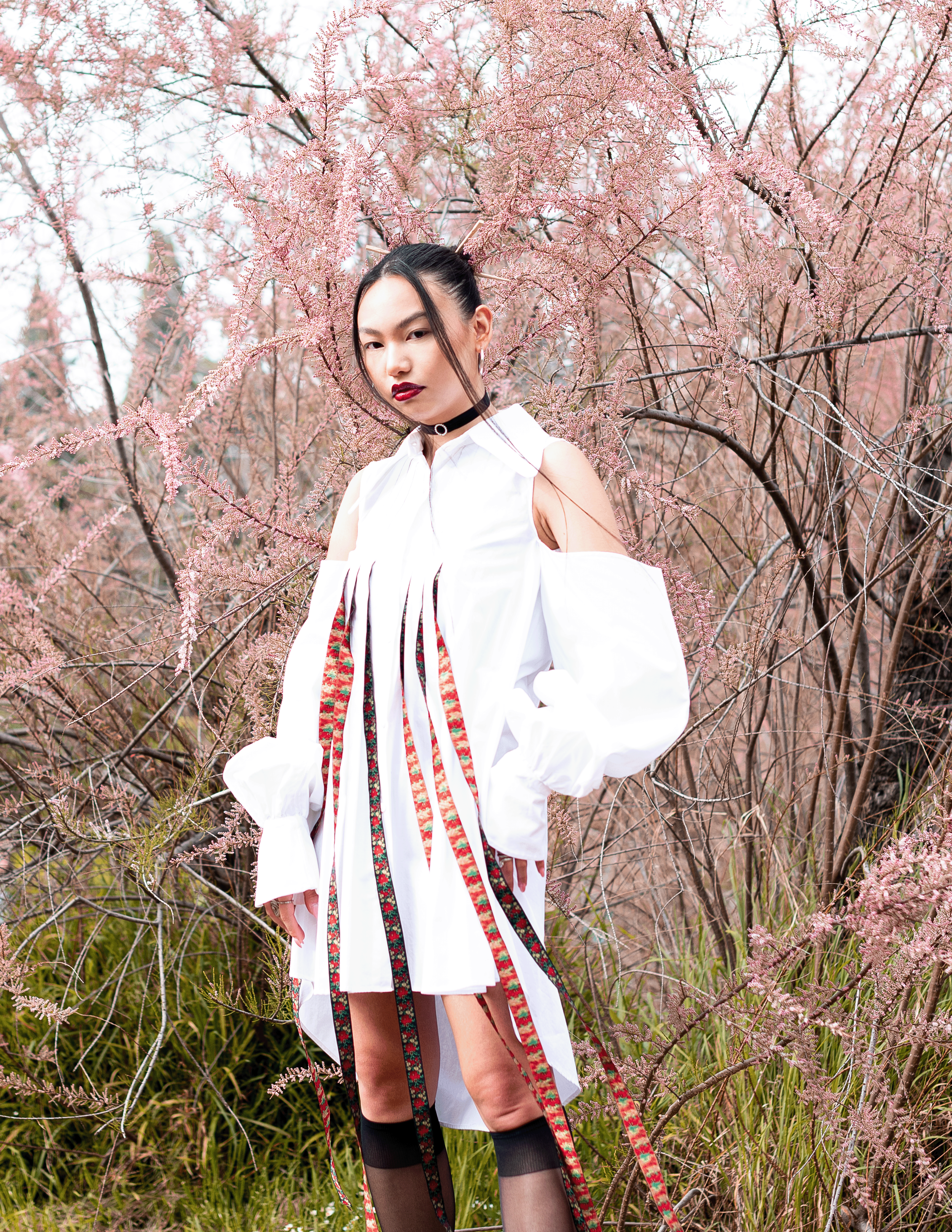 Una donna vestita con un abito bianco corto e calze nere trasparenti si trova tra cespugli di fiori rosa primaverili, con capelli raccolti e trucco scuro.